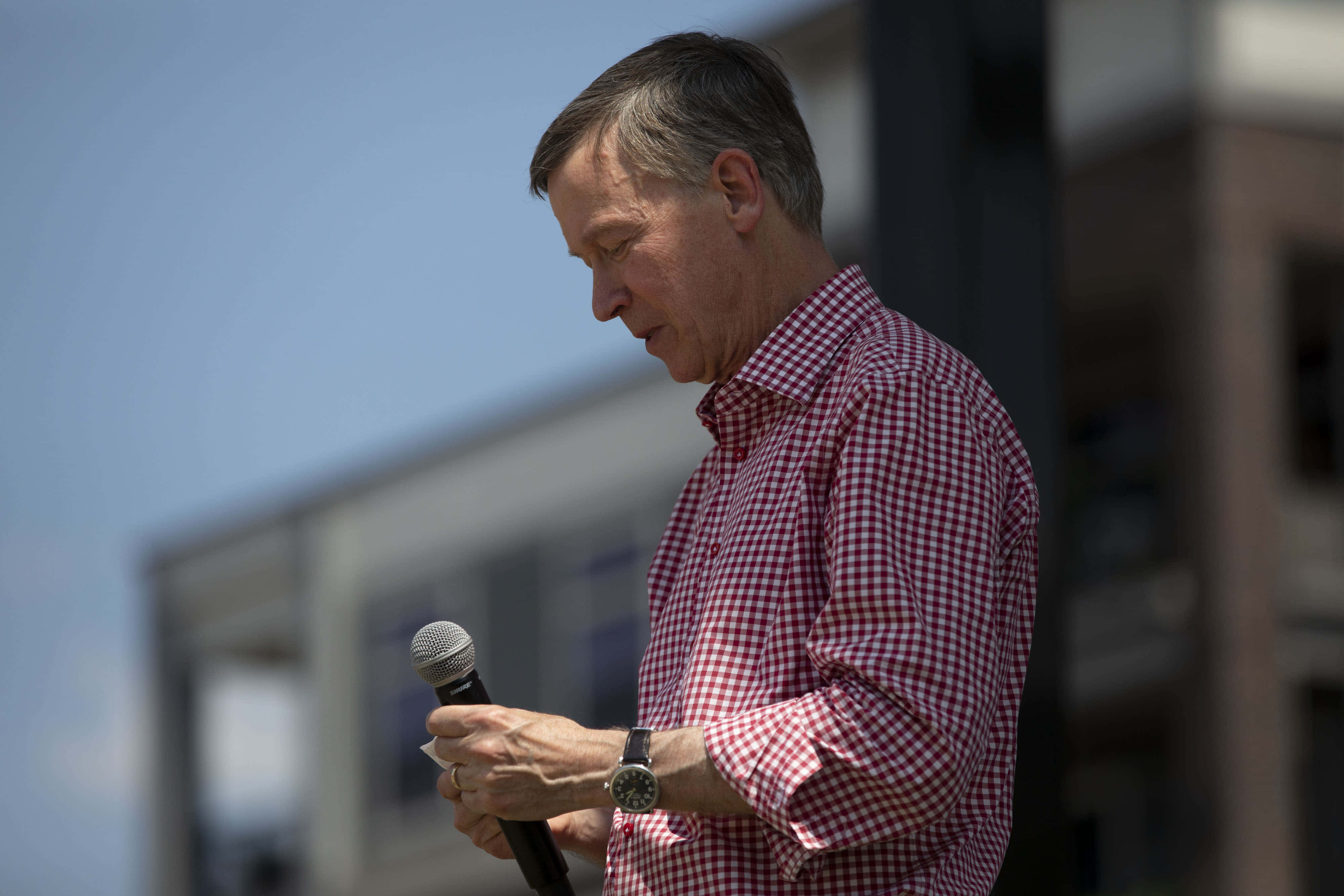 John Hickenlooper addressing audience with Microphone 壁紙 — 一名身穿紅白格仔恤衫的男子在晴朗藍天下於戶外對著咪高峰說話。
