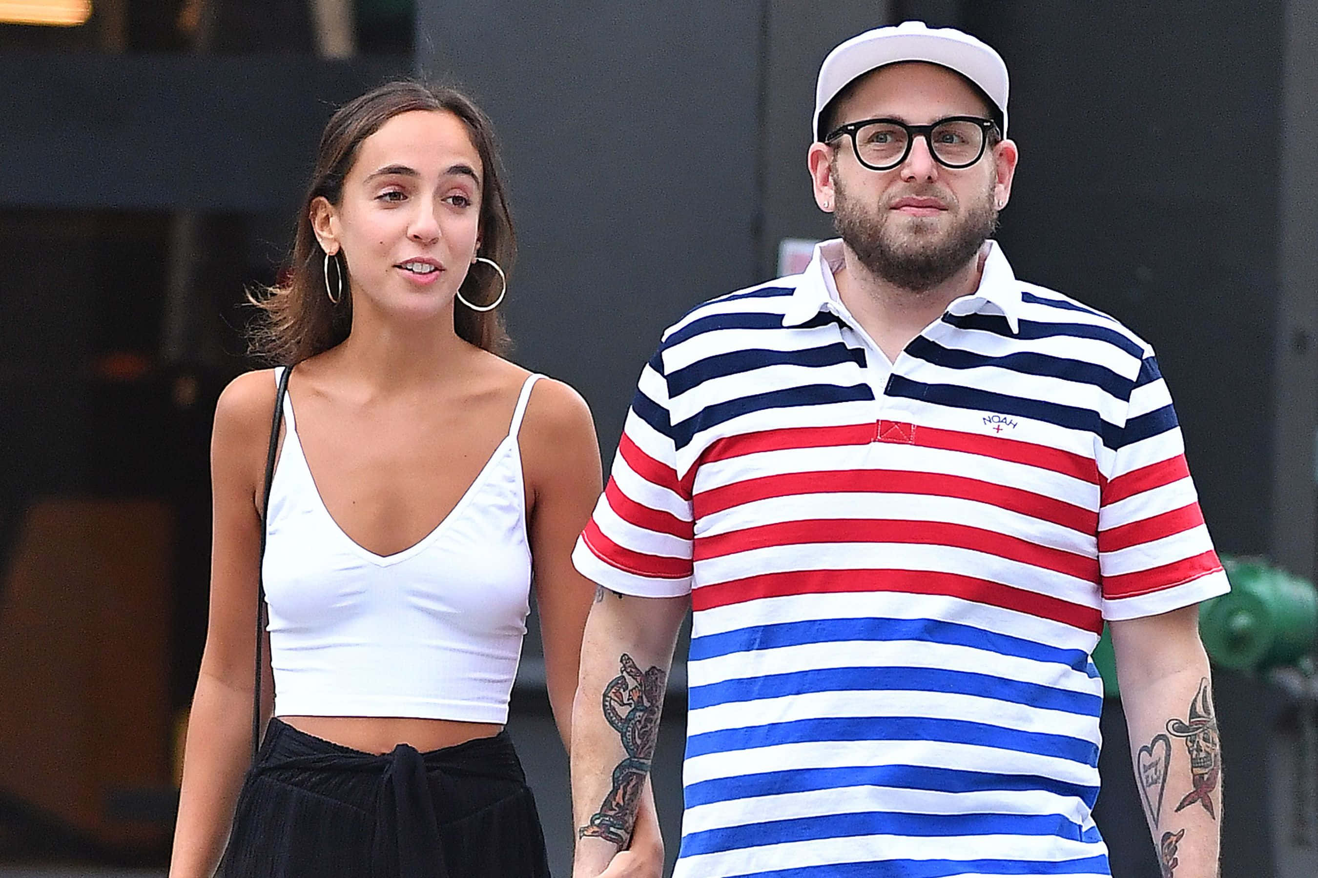 jonah hill 桌布 壁紙 — 一名身穿紅白藍條紋Polo衫的男子和一名身穿白色露臍上衣的女子手牽手在戶外散步。