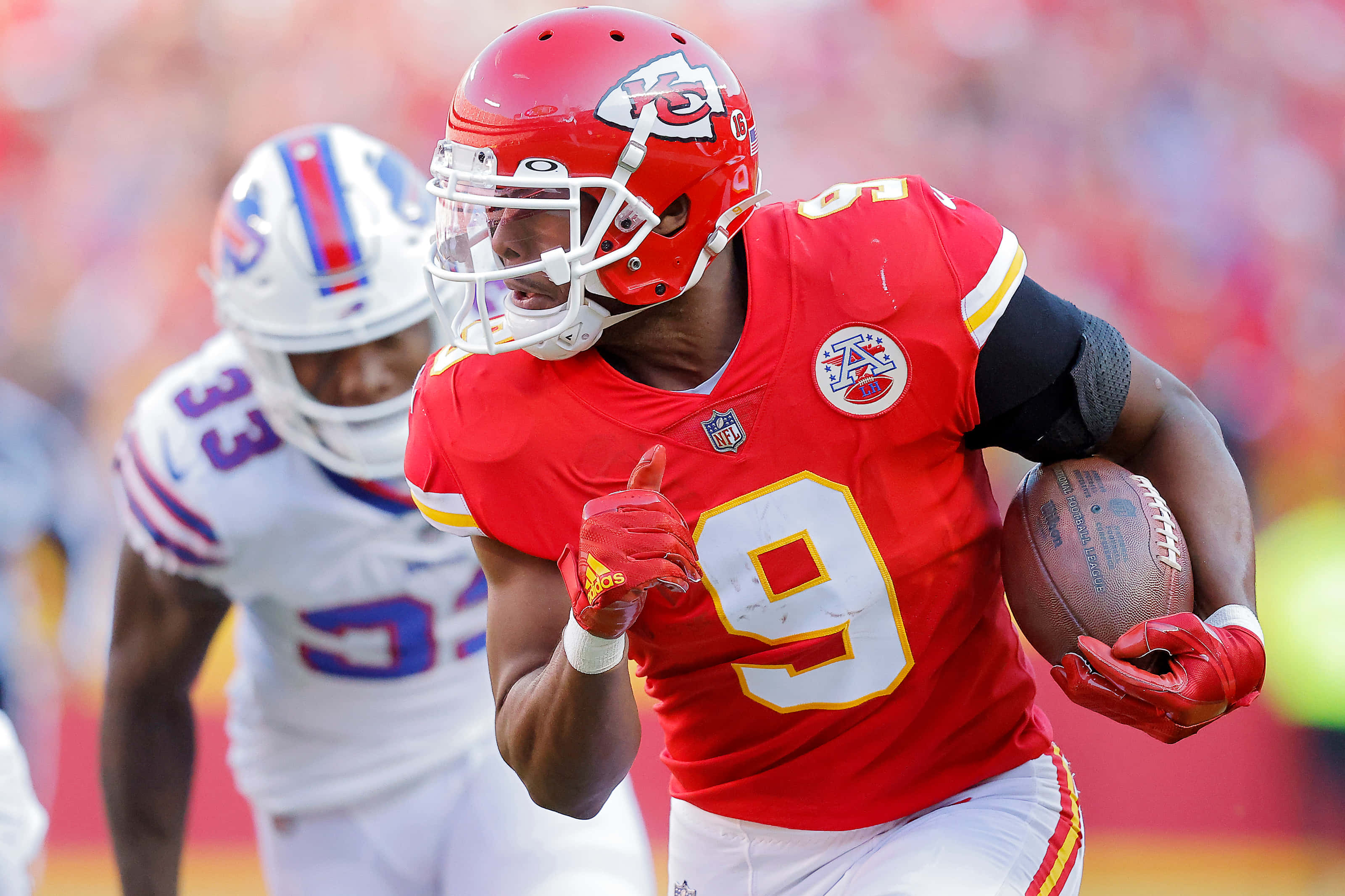 Juju Smith Schuster 桌布 壁紙 — 一名身穿紅色9號球衣的堪薩斯城酋長隊美式足球員在日間比賽中帶球奔跑，身後有一位模糊的白色對手。