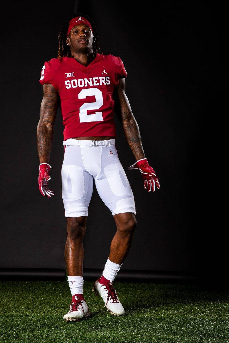 Sooners Ceedee Lamb 壁紙 — 一名身穿紅色Sooners球衣二號的美式足球員自信地站在黑色背景前的人造草皮上。