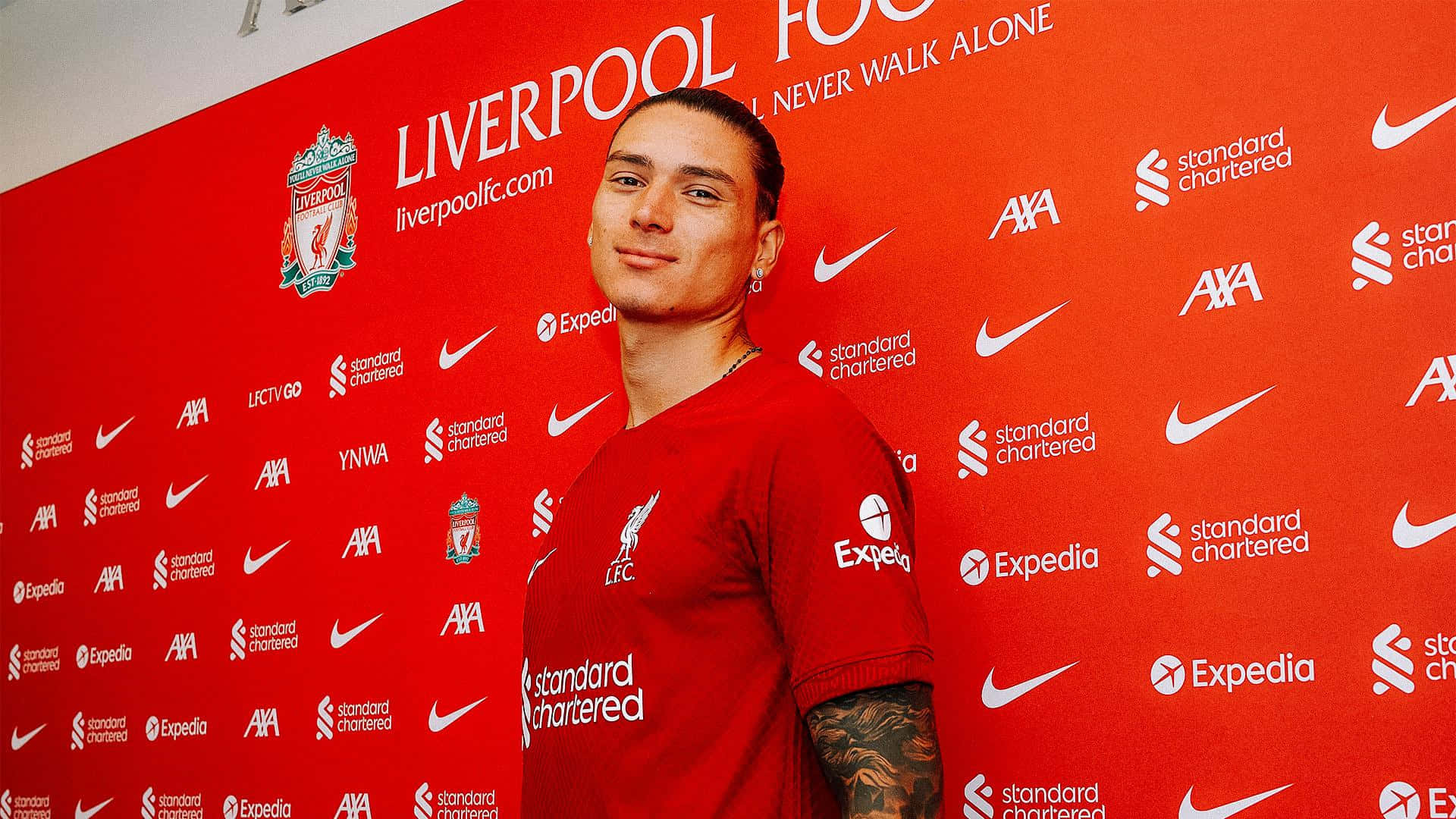 Liverpool 桌布 壁紙 — 一名身穿紅色利物浦球衣的足球員，在充滿贊助商標誌的鮮紅色品牌牆前自信地擺姿勢。