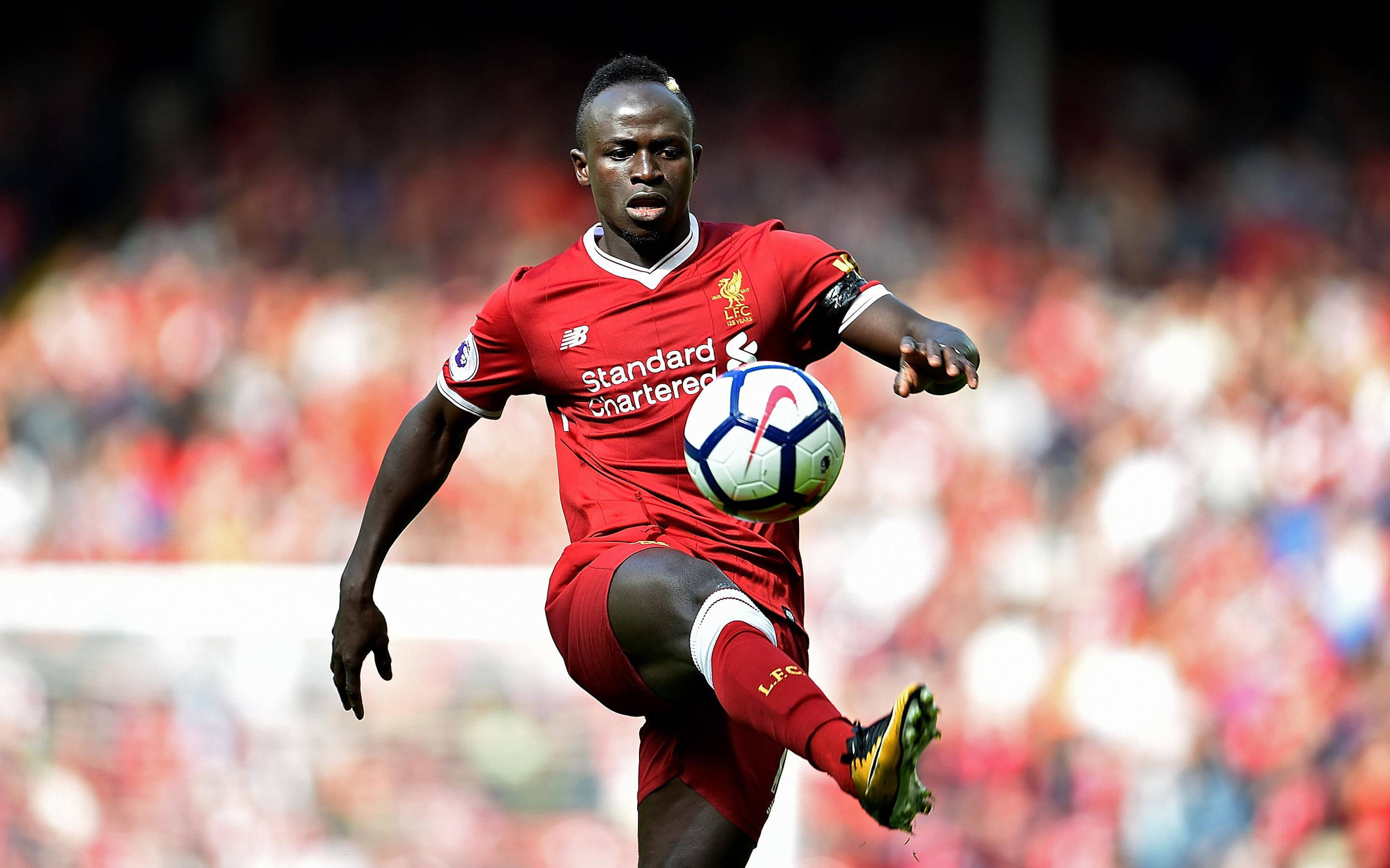 Liverpool Fc 桌布 壁紙 — 一名身穿紅色利物浦球衣的足球運動員在陽光明媚的球場上，在跨步中控制著一個白藍相間的球，身後是模糊的體育場人群。