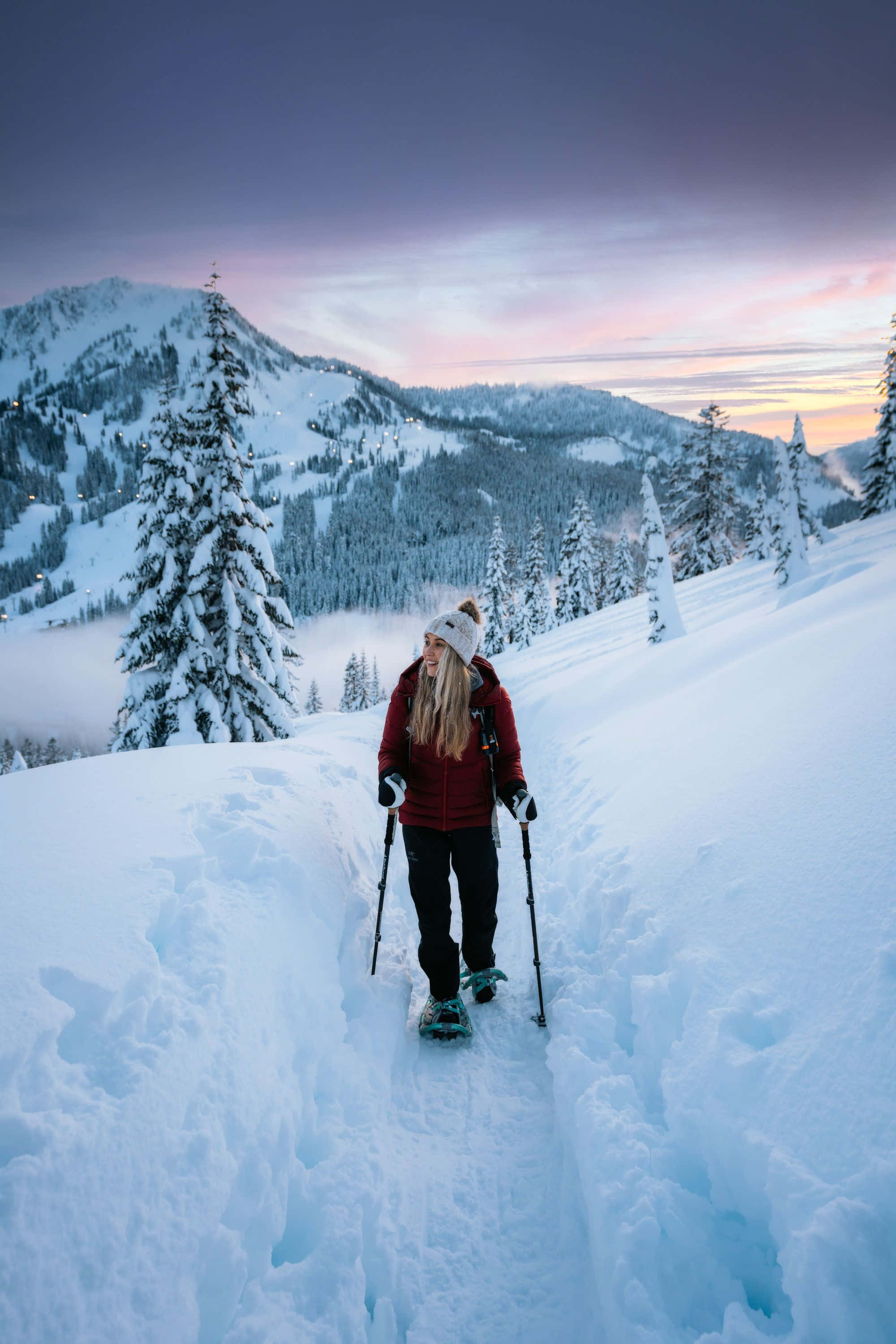 Winter Hiking 桌布 壁紙 — 一名身穿紅色外套的女子在黃昏時分，在白雪覆蓋的松樹和紫粉色天空下，徒步穿越深雪覆蓋的山間小徑。