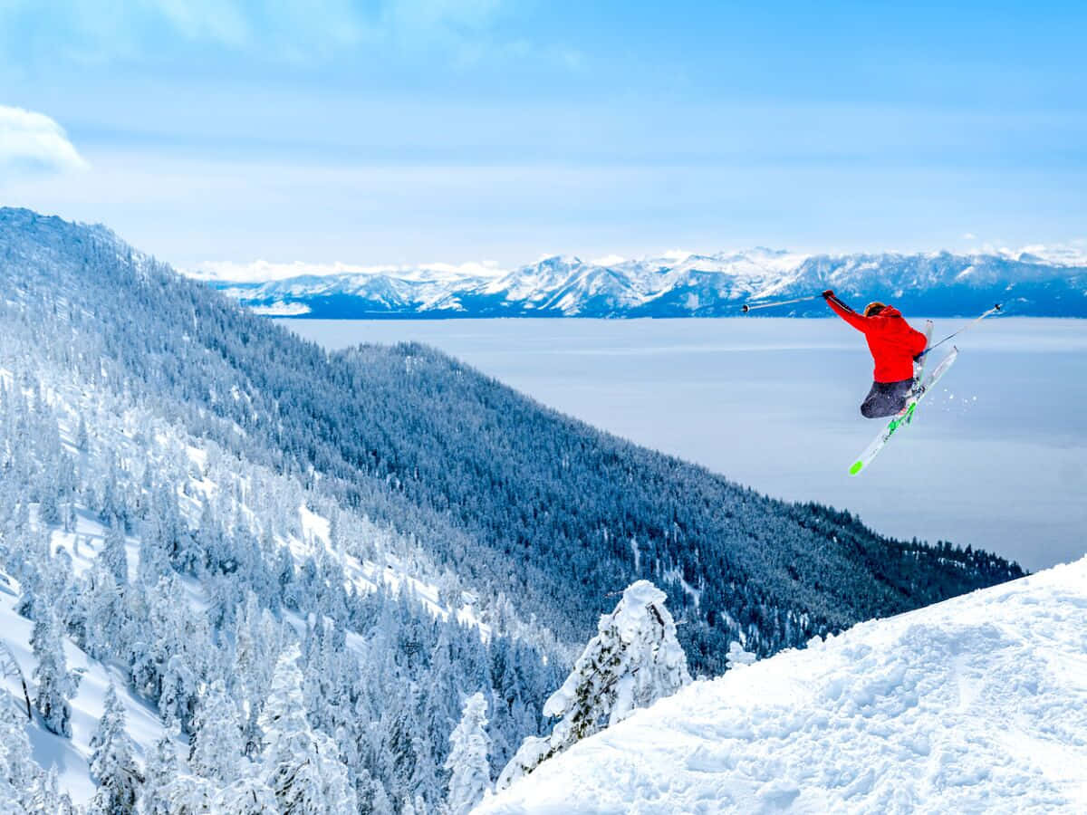 Skiing 桌布 壁紙 — 一名身穿紅色外套的滑雪者在晴朗的藍天下，飛越俯瞰冰凍湖泊的雪山斜坡。