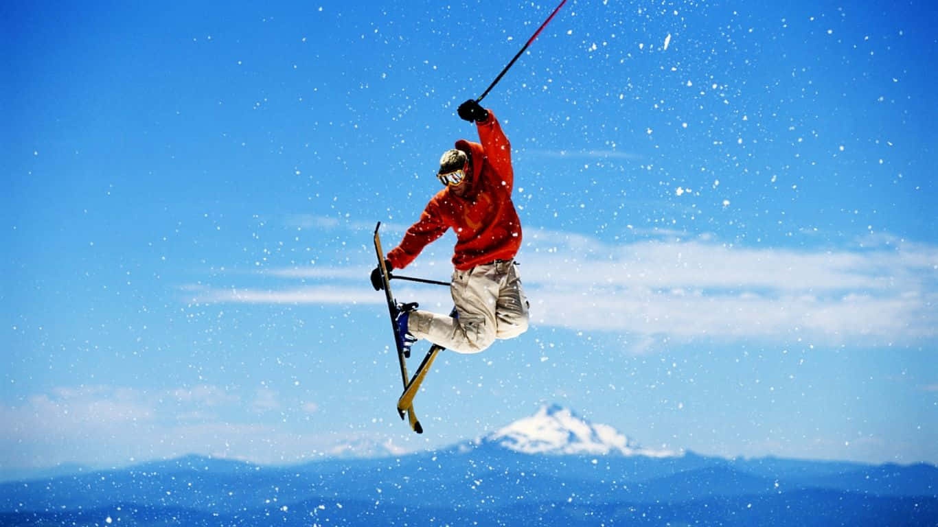 Winter Sports 桌布 壁紙 — 一名身穿紅色外套的滑雪者在雪空中飛翔，背景是明亮的藍天和遠處的雪山。