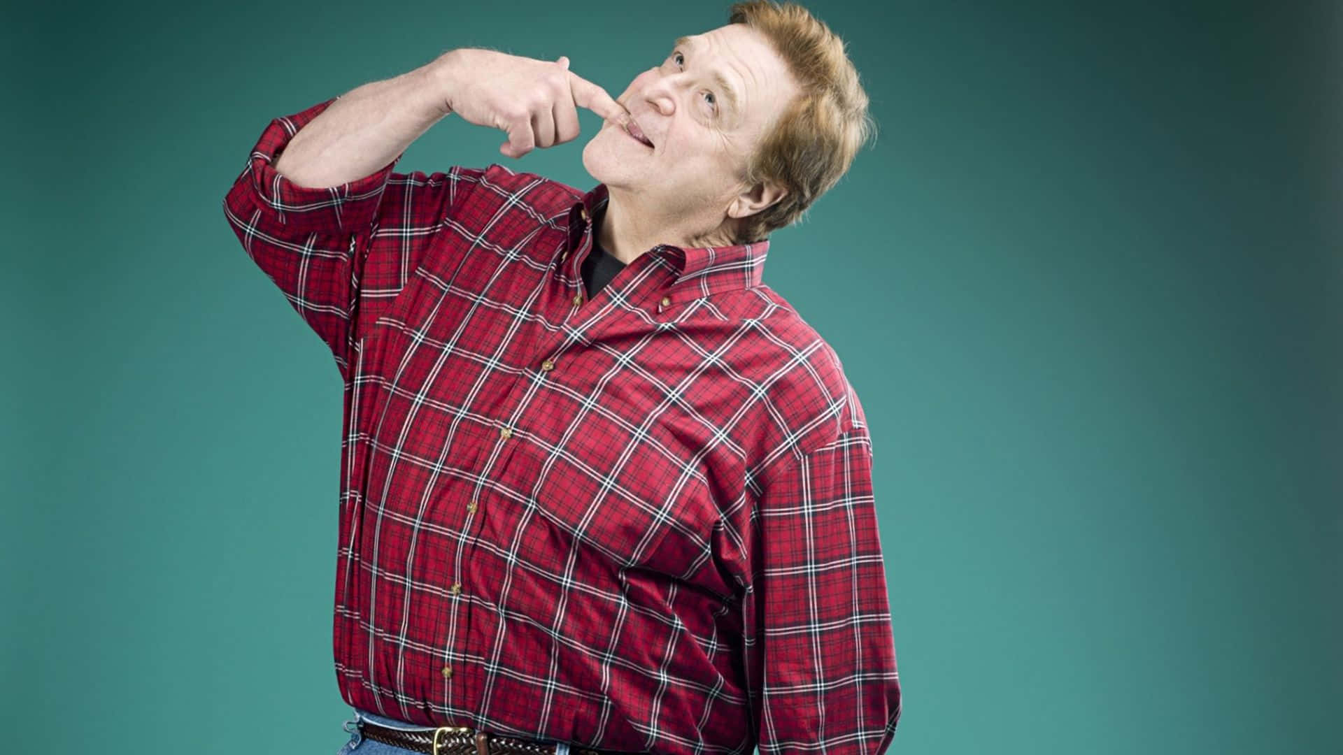 John Goodman 桌布 壁紙 — 一名身穿紅色格仔恤衫的男子，在青綠色工作室背景前，帶著俏皮表情指著自己的鼻子。