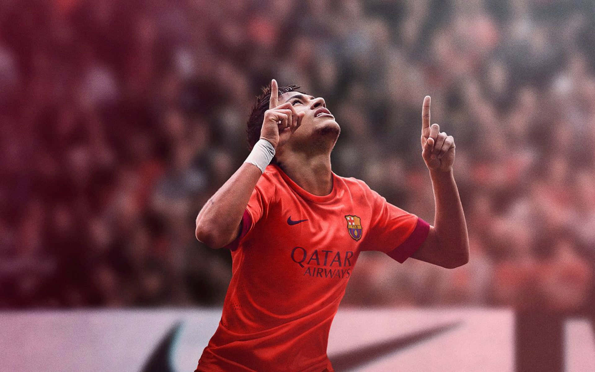 Neymar 桌布 壁紙 — 一名身穿紅色球衣的足球運動員在模糊的體育場人群中，雙手指向天空，勝利地慶祝。