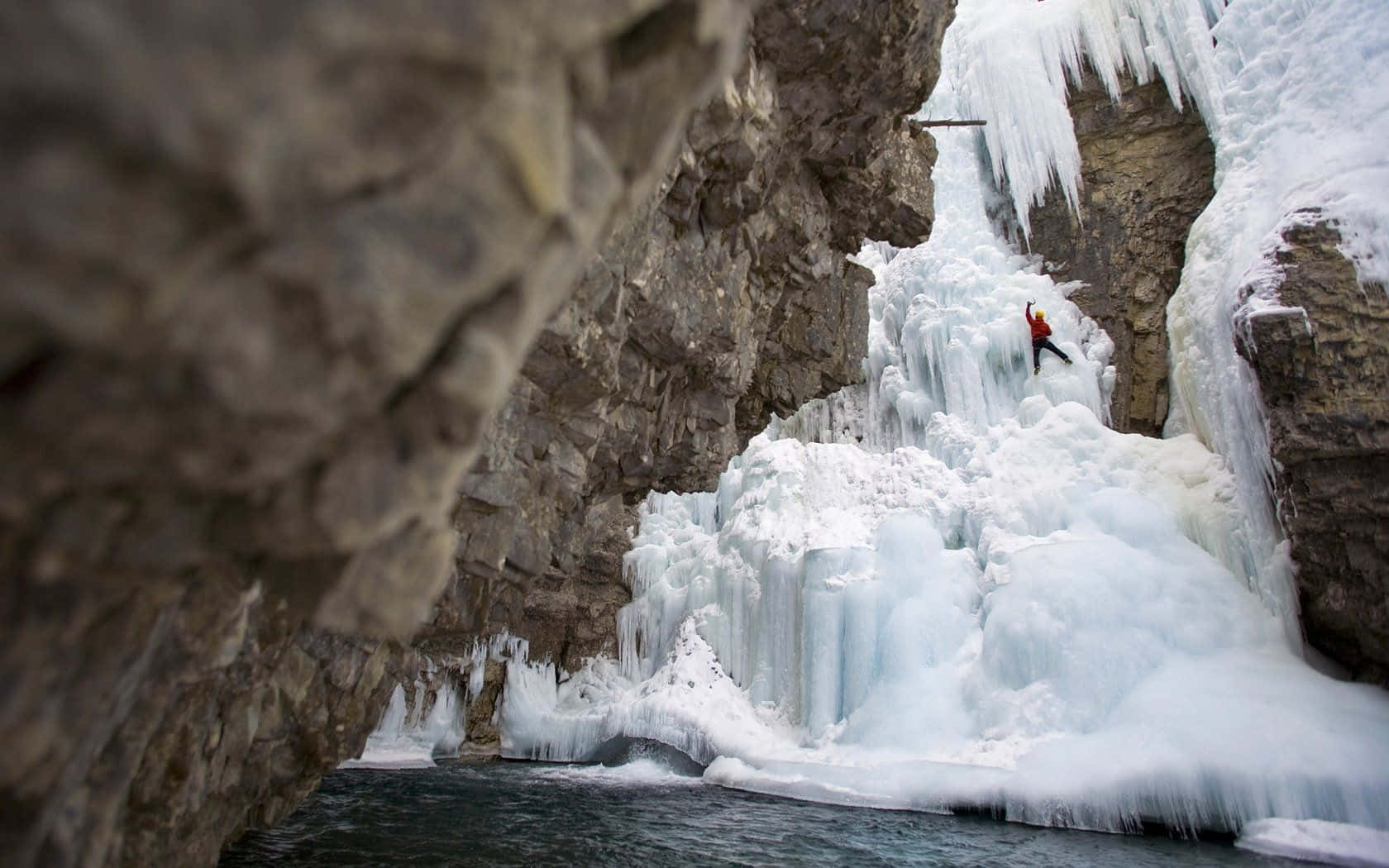 Ice Climbing 桌布 壁紙 — 一名身穿紅色衣服的攀岩者正在攀登一座高聳的冰凍瀑布，周圍環繞著崎嶇的懸崖和冰冷的冰柱。