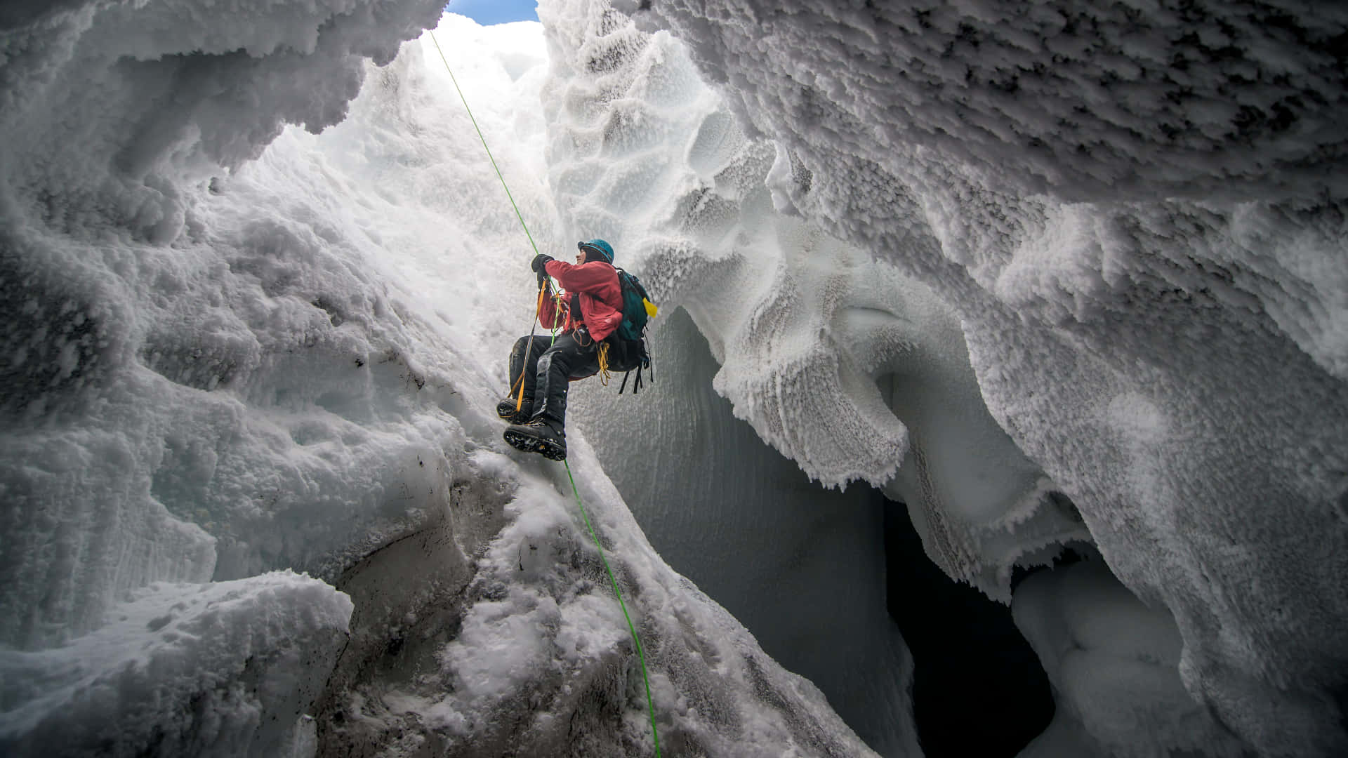 Ice Climbing 桌布 壁紙 — 一名身穿紅色裝備的攀冰者，在冰凍的冰川洞穴內，使用冰斧和綠色繩索攀登高聳的冰壁。