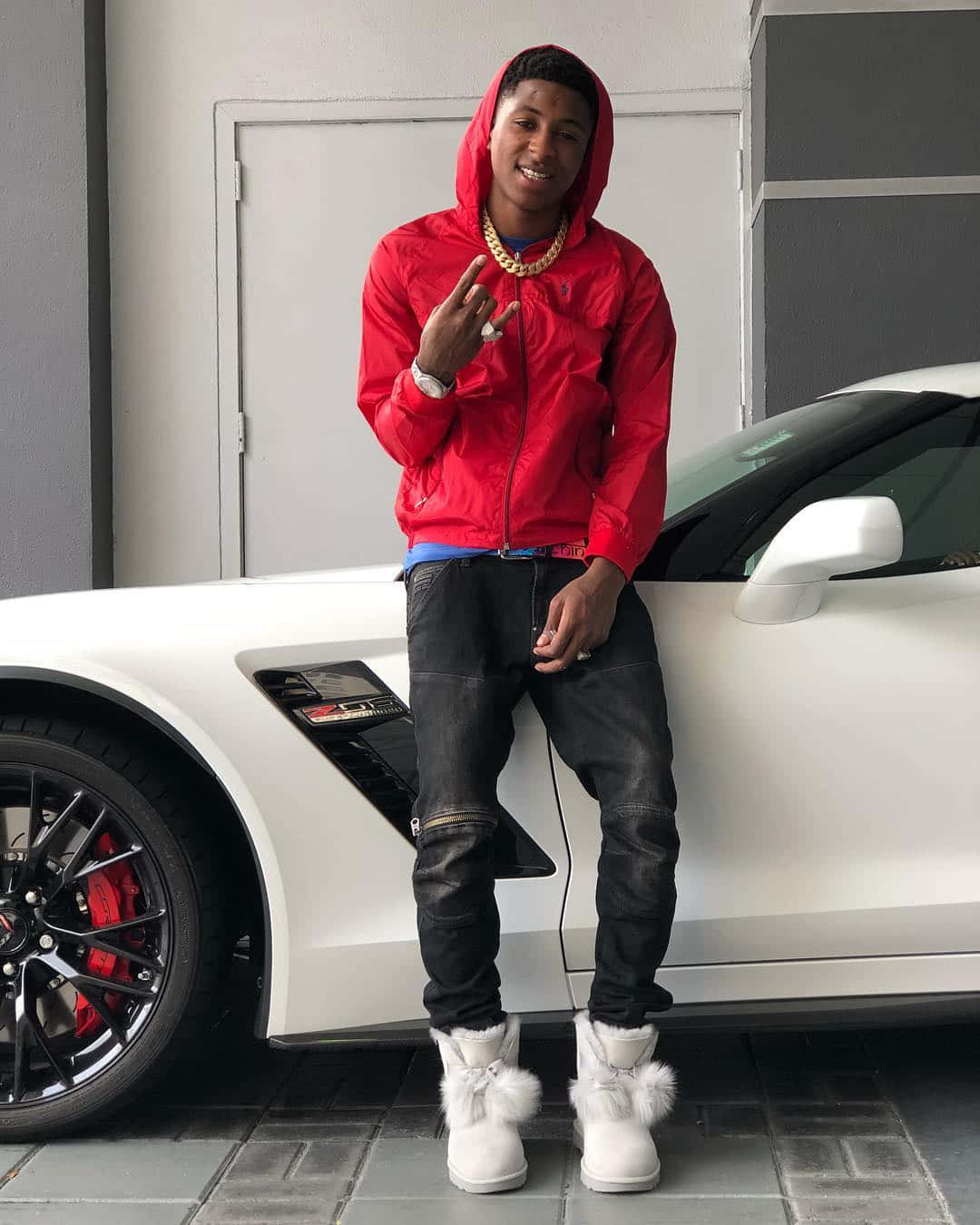 NBA Youngboy 壁紙 — 一名身穿紅色連帽衫和白色毛靴的年輕男子在車庫裡自信地站在一輛白色跑車旁邊。