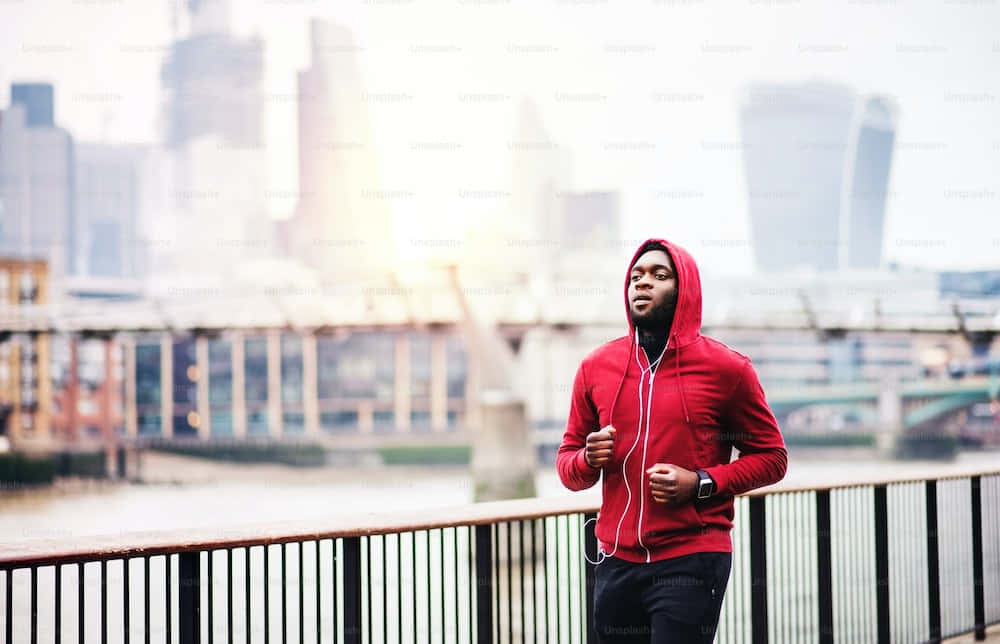 Red Hoodie 桌布 壁紙 — 一名身穿紅色連帽衫的男子沿著河邊欄杆奔跑，背景是朦朧的城市天際線。