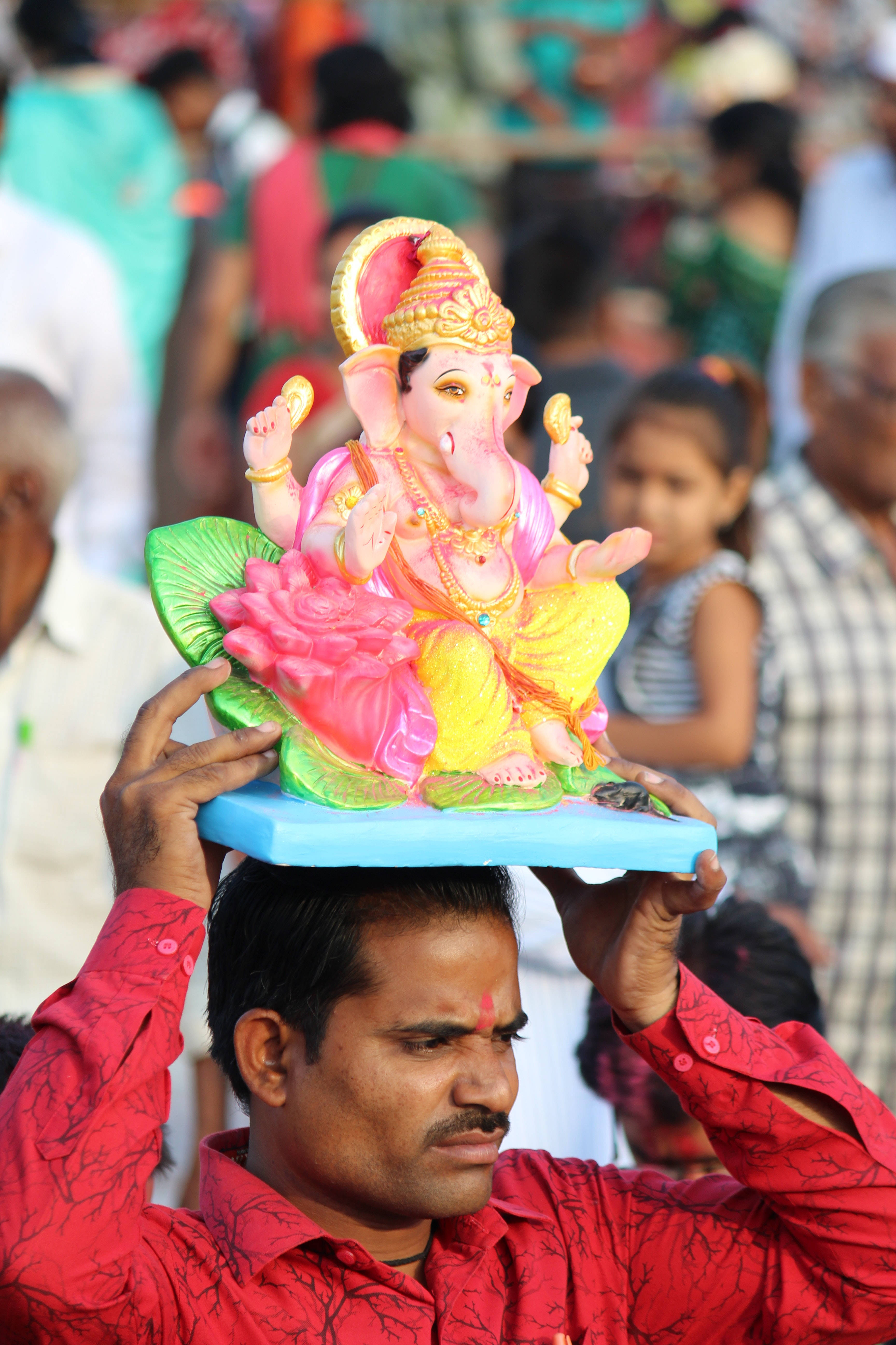 Ganesh Chaturthi 桌布 壁紙 — 一名身穿紅衫的男子在擁擠的節日場景中，將色彩繽紛的象神神像平衡在頭頂上。