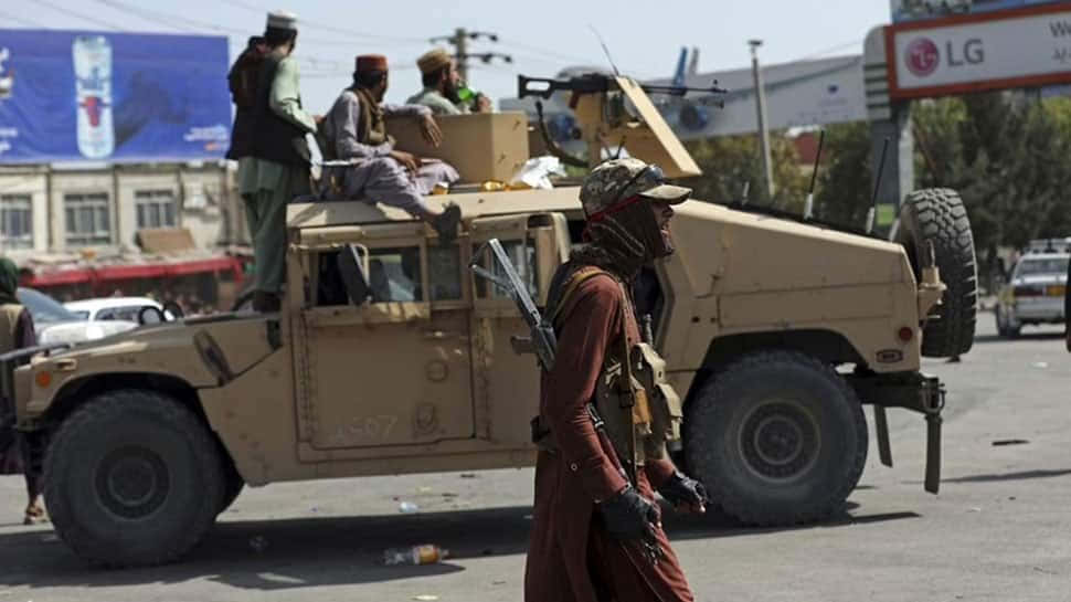 Taliban 桌布 壁紙 — 一名身穿紅袍的士兵走過一輛棕褐色裝甲車，車上有武裝人員，在塵土飛揚的城市街道上。