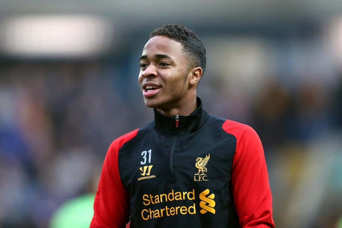 Sterling 桌布 壁紙 — 一名身穿紅黑利物浦訓練服的年輕足球員站在模糊的球場上微笑。
