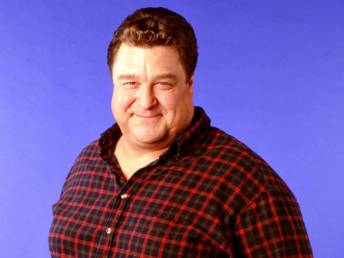 John Goodman 桌布 壁紙 — 一名身穿紅黑格仔恤衫嘅男士喺純藍色攝影棚背景前溫暖咁微笑。