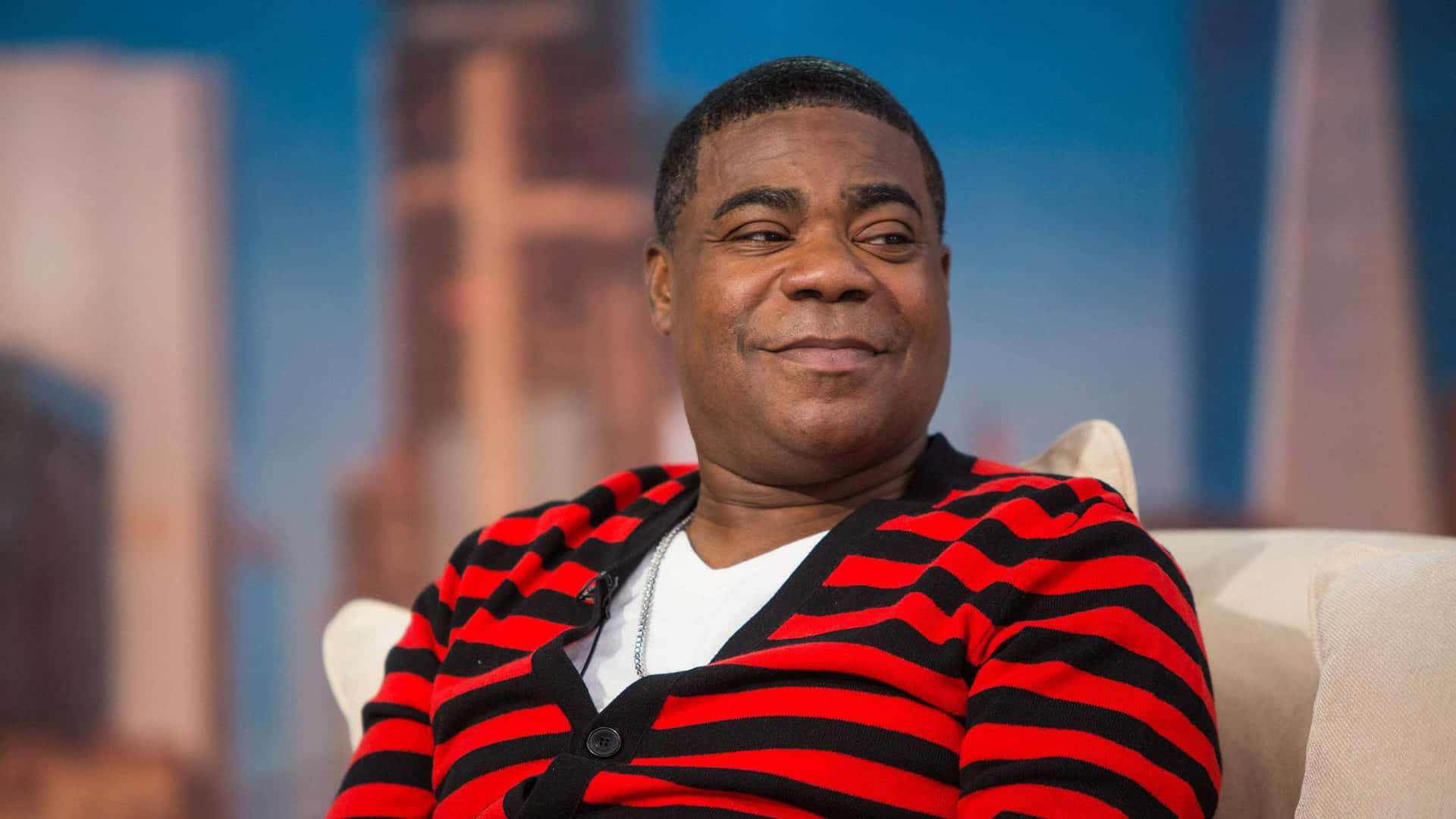 Tracy Morgan 桌布 壁紙 — 一名身穿紅黑間條開襟毛衣的男子坐在白色椅子上，對著模糊的城市景觀背景露出溫暖的笑容。
