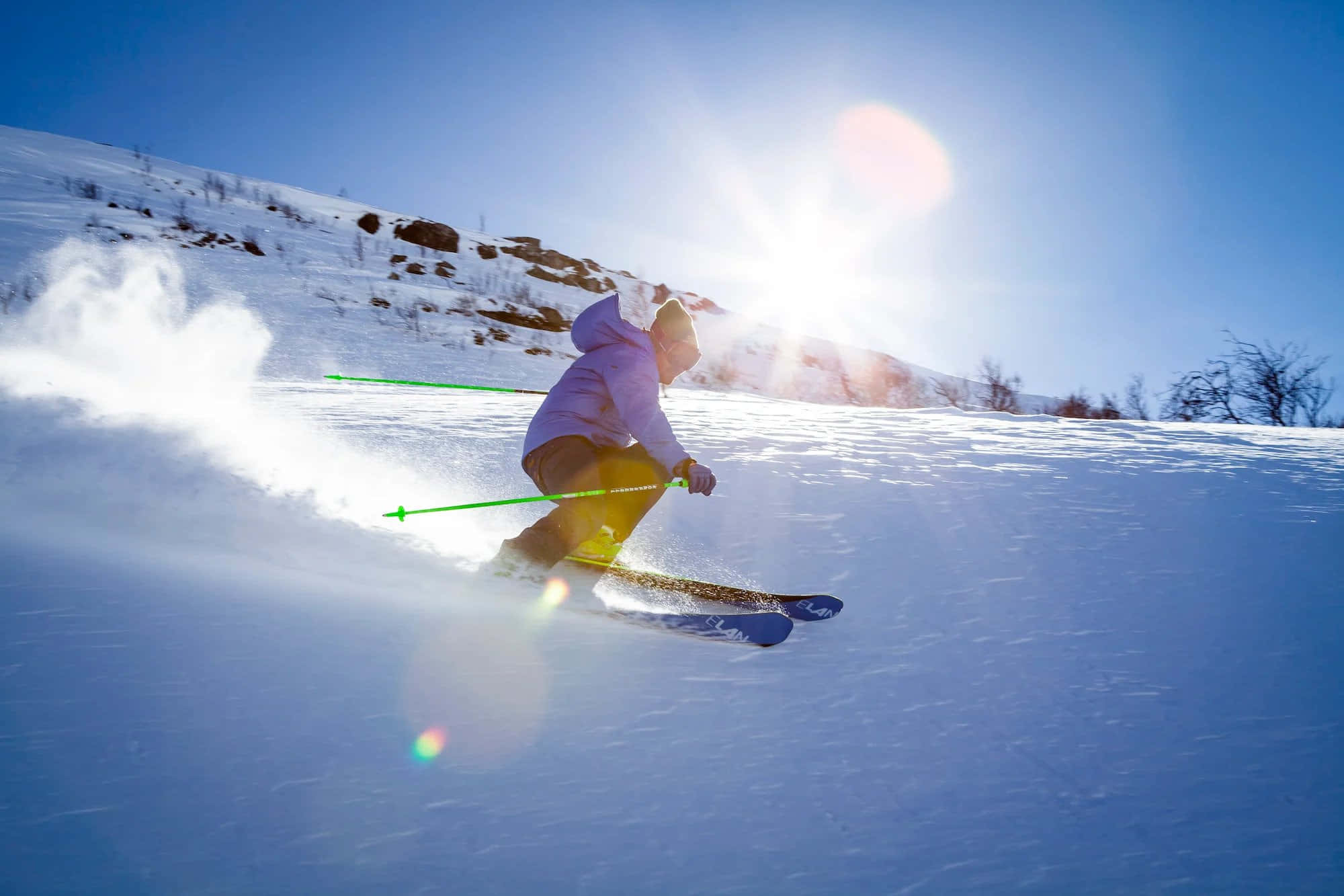 Skiing 桌布 壁紙 — 一名身穿紫色外套的滑雪者，在陽光普照、帶有鏡頭眩光的斜坡上，劃過新雪。