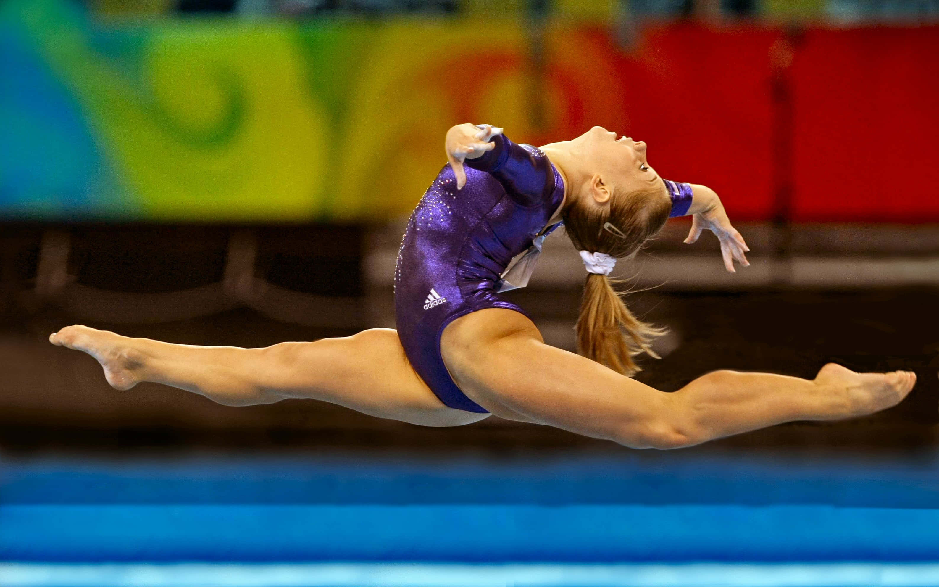 Gymnastics 桌布 壁紙 — 一名身穿紫色緊身衣的女體操運動員在比賽套路中於空中執行動態的劈叉跳躍。