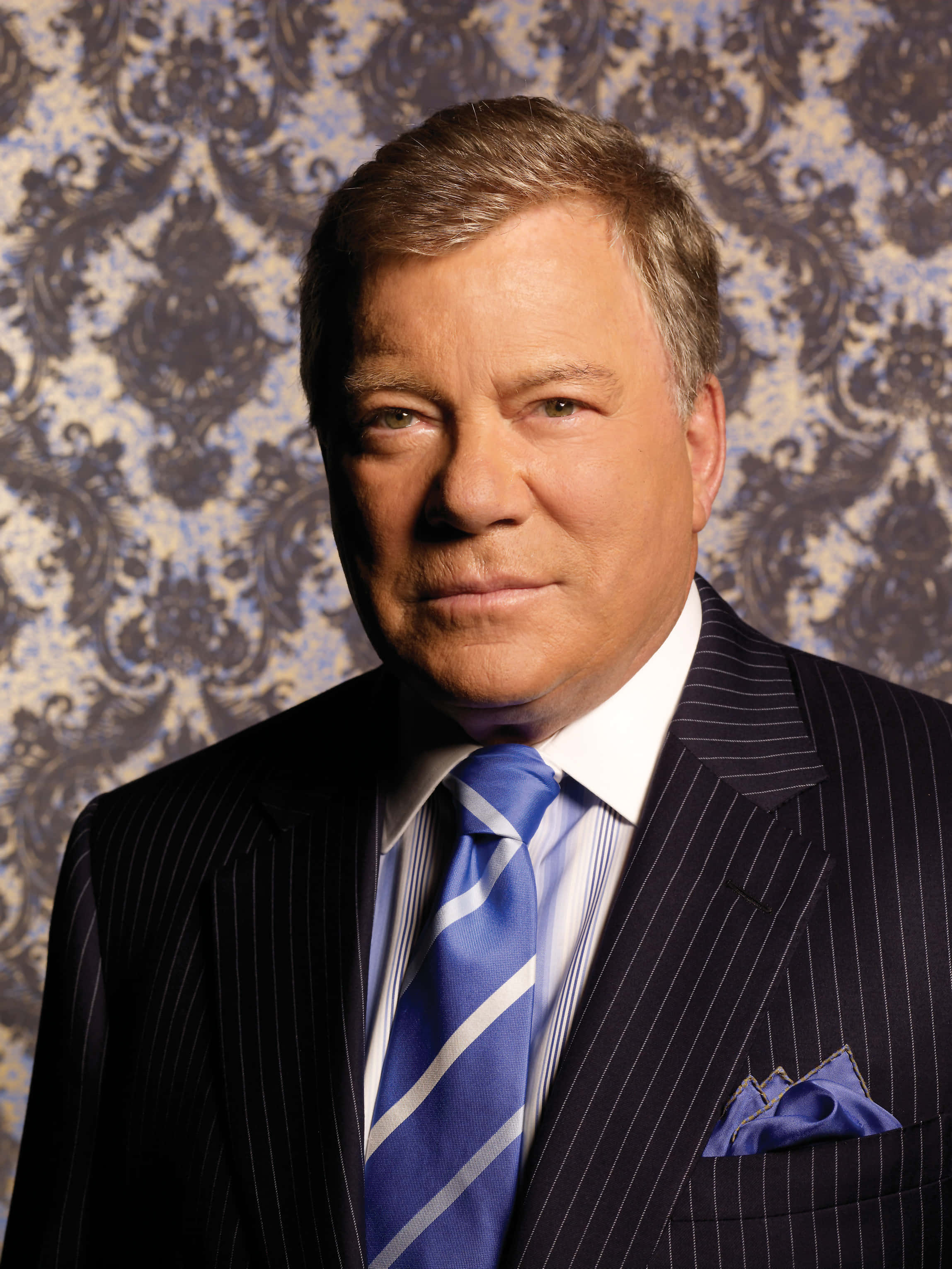 william shatner 桌布 壁紙 — 一名身穿細條紋西裝和藍色領帶的男子，表情嚴肅地站在華麗的錦緞牆紙前。