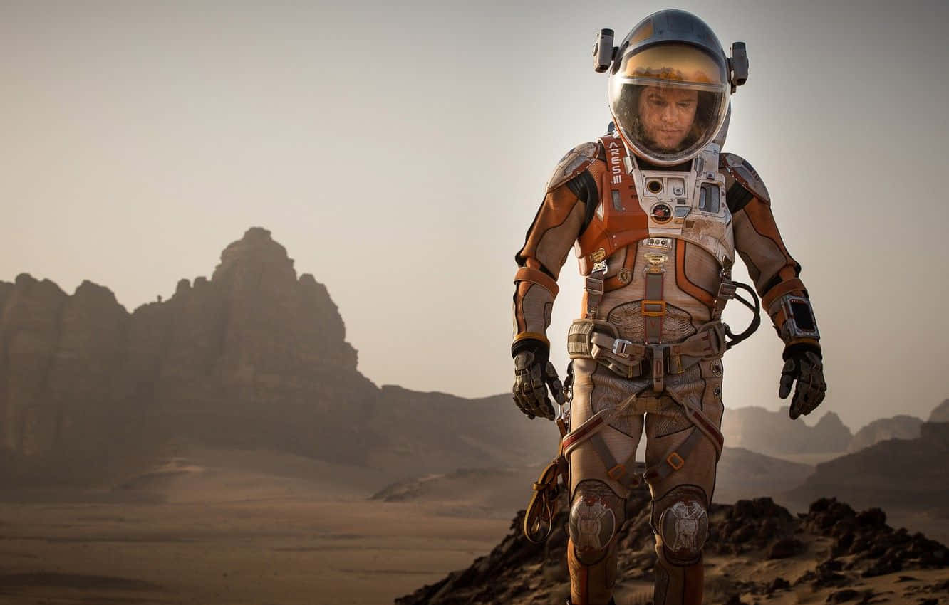 The Martian 桌布 壁紙 — 一名身穿細節豐富的橙色和白色太空衣的太空人在朦朧天空下，走過一個有遠處平頂山的荒蕪岩石行星。