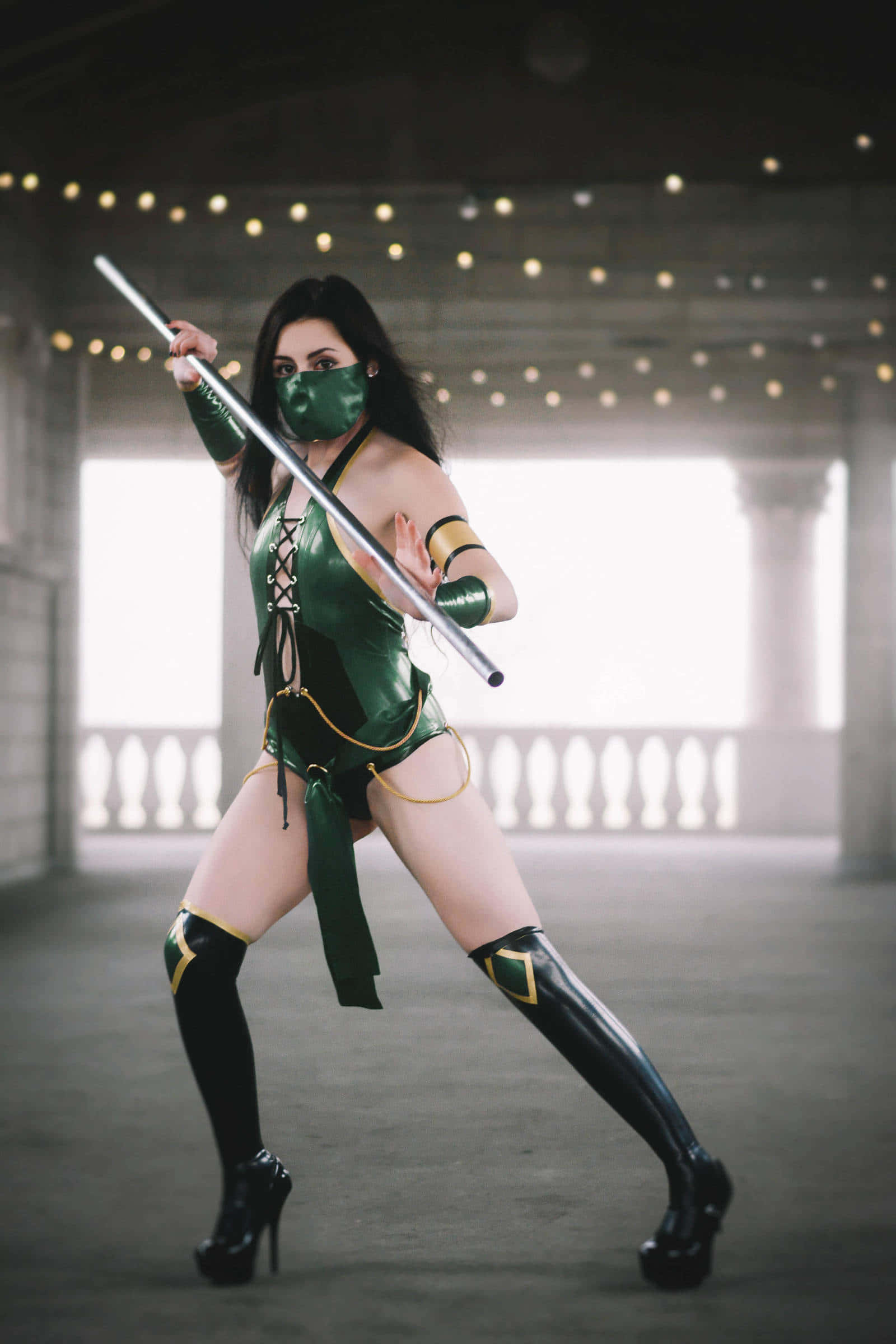 Mortal Kombat Cosplays 桌布 壁紙 — 一名身穿綠色和金色忍者服裝的女性，在燈串下以戲劇性的姿勢揮舞著棍棒。