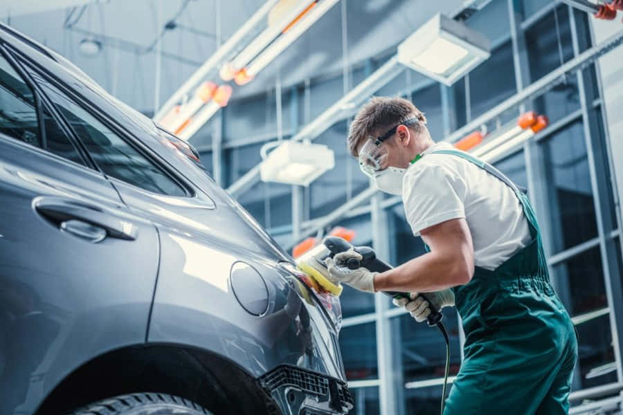 Car Repair 桌布 壁紙 — 一名身穿綠色工裝褲和安全裝備的機械師在現代汽車美容車庫中仔細打磨一輛光滑的灰色汽車。