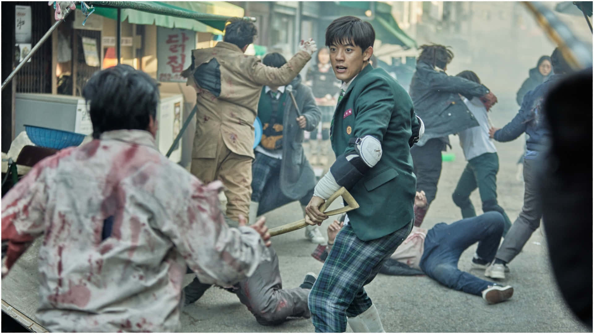 All Of US Are Dead 桌布 壁紙 — 一名身穿綠色校服的年輕人在混亂中揮舞著鏟子，人們在煙霧瀰漫的城市街道上打鬥流血。