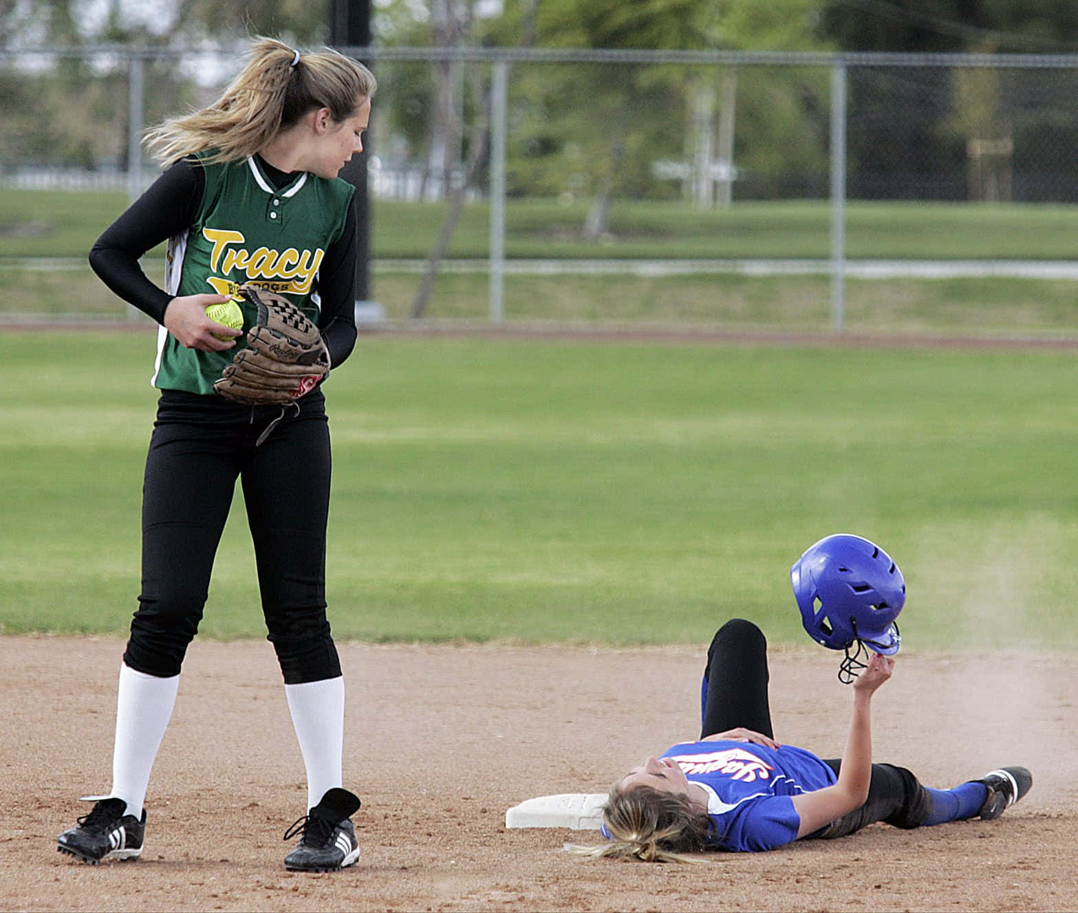 Softball 桌布 壁紙 — 一名身穿綠色球衣的壘球手站立持球，另一名身穿藍色球衣的隊友躺在泥地壘道上抱著頭盔。