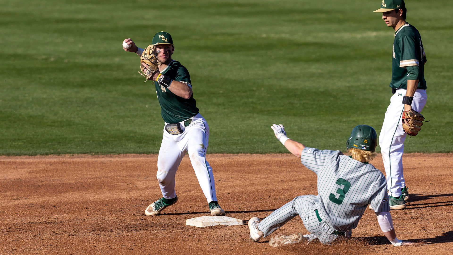 College Baseball 桌布 壁紙 — 一名身穿綠色球衣的棒球手擲向一壘，同時一名身穿灰色球衣的跑壘員在陽光明媚的日間比賽中滑入壘包。