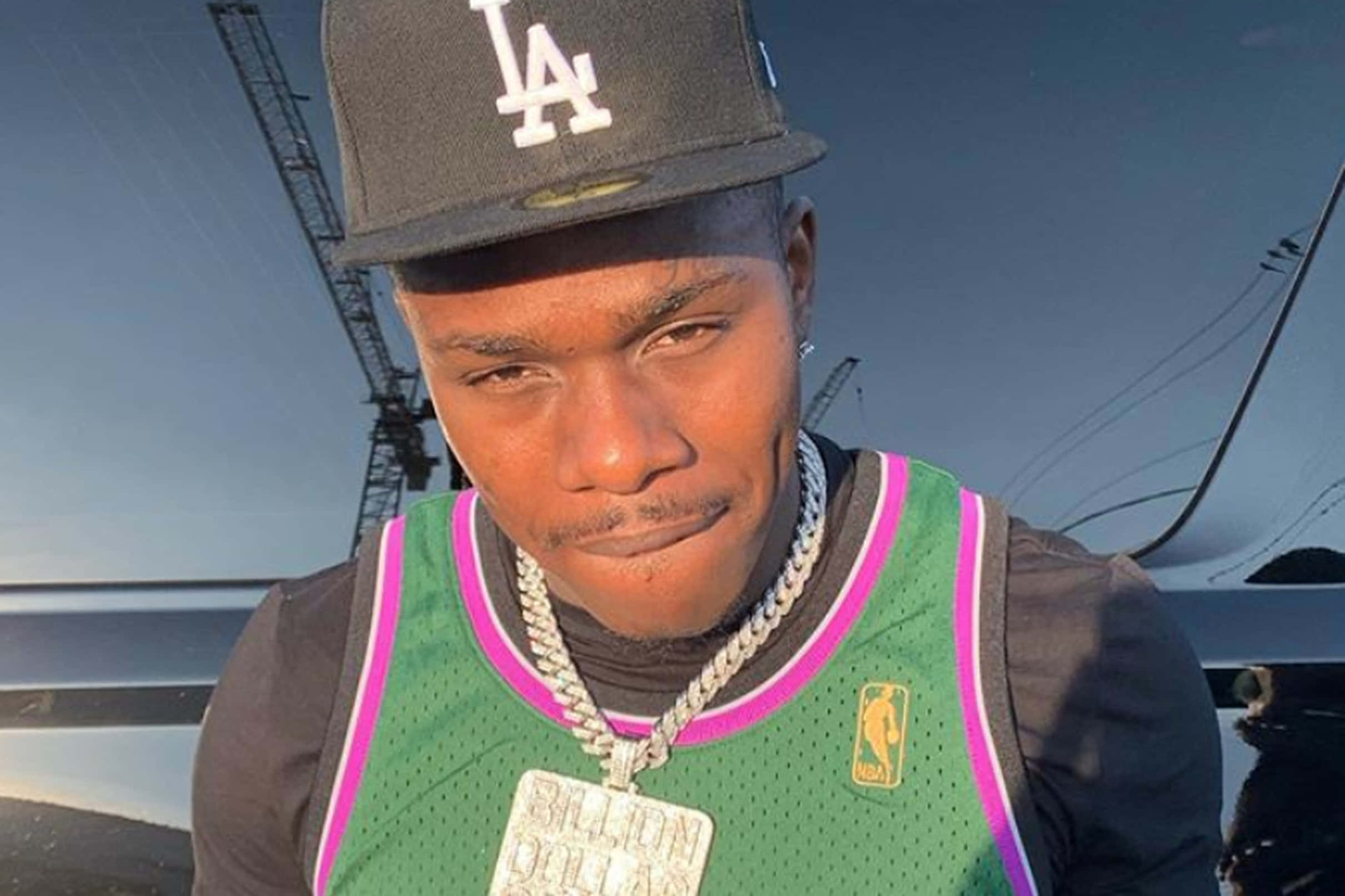 Dababy 桌布 壁紙 — 一名身穿綠色籃球球衣和LA帽子的男子，自信地戴著一條大鏈條，以汽車和建築起重機為背景擺姿勢。