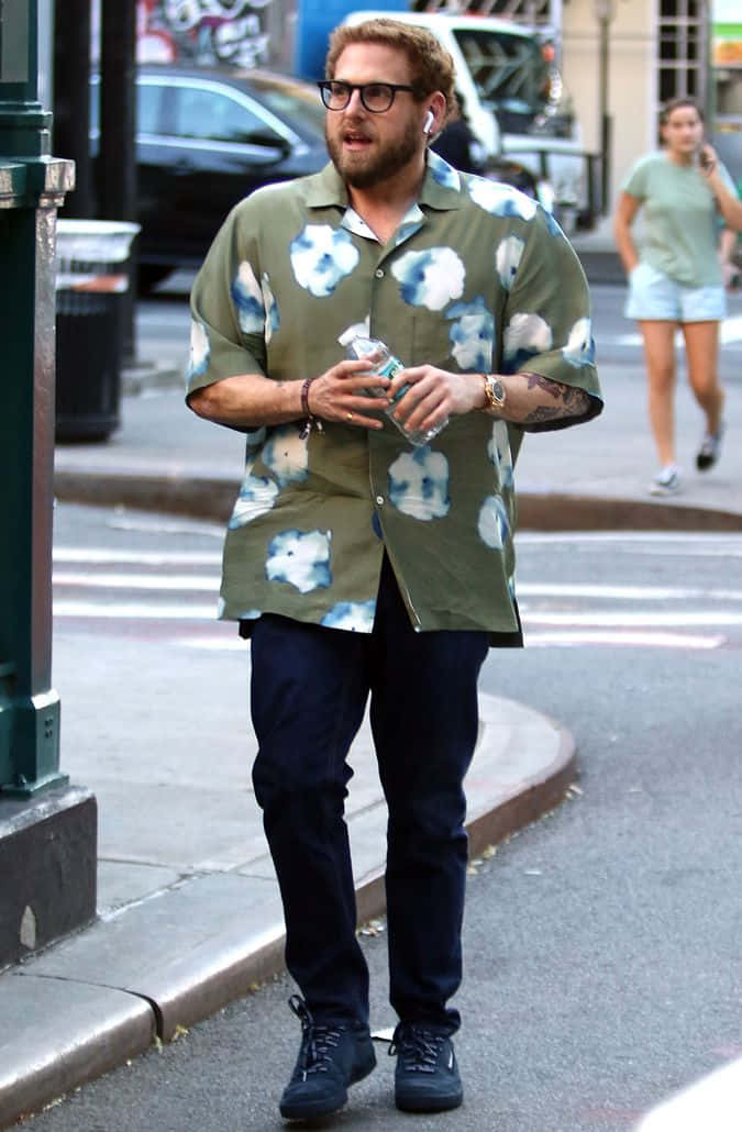 jonah hill 桌布 壁紙 — 一名身穿綠色花卉圖案襯衫的男子，戴著耳塞，拿著水樽，在城市行人路上行走。
