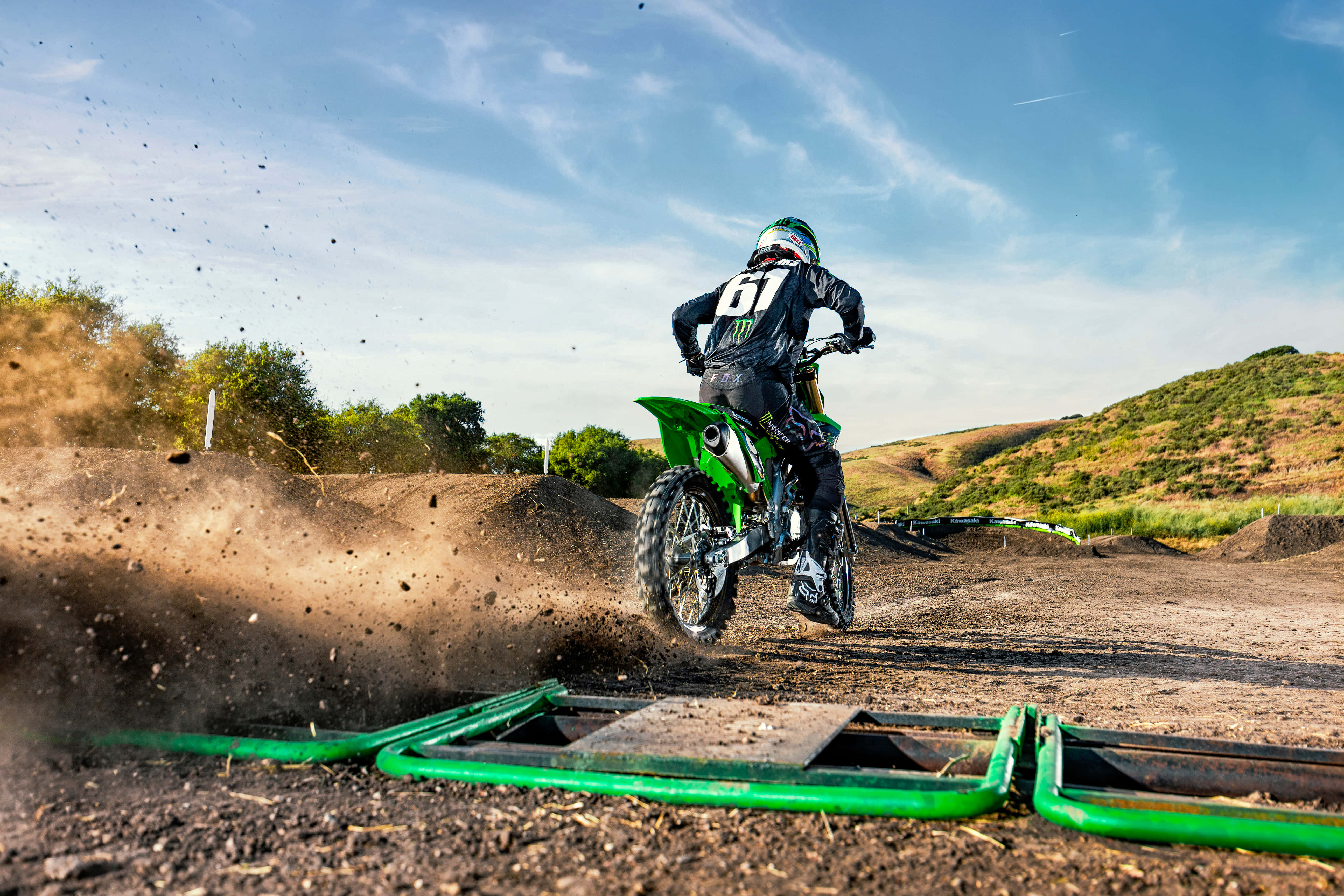 Dirtbike 桌布 壁紙 — 一名身穿綠色裝備的越野電單車手在陽光明媚的賽道上飛越泥土跳台，塵土飛揚，背景是山丘。