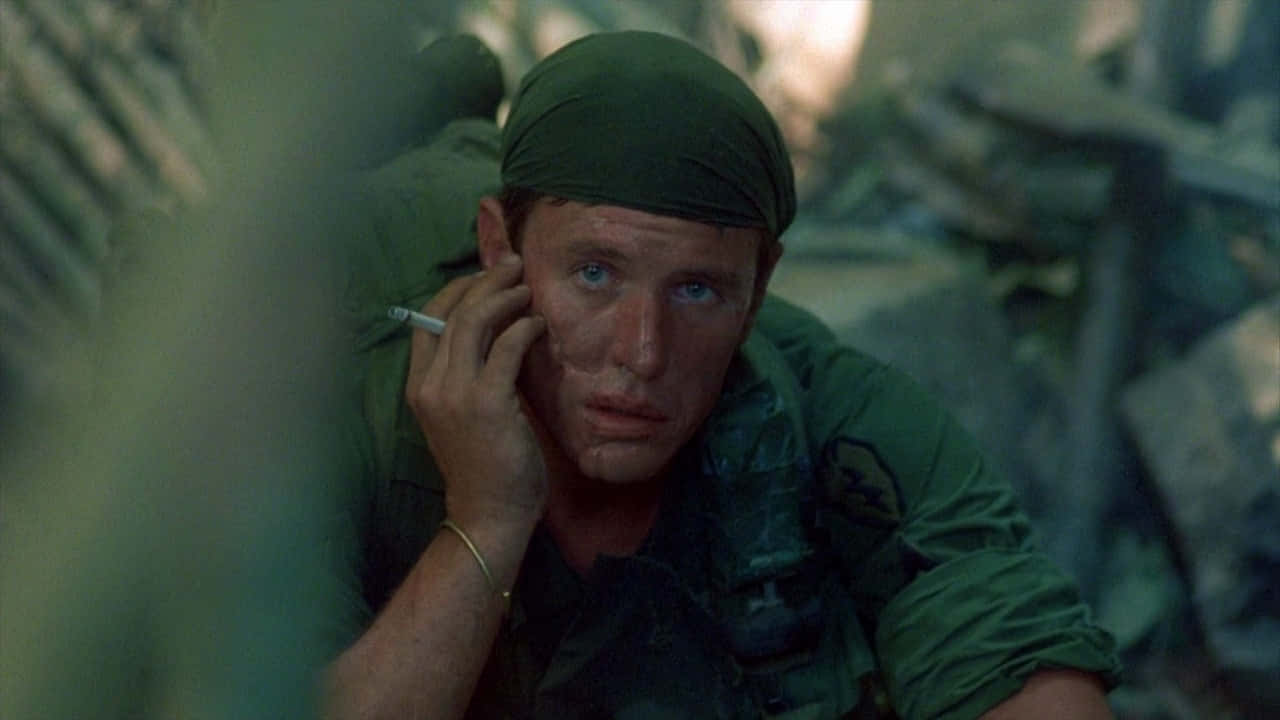 Tom Berenger 桌布 壁紙 — 一名身穿綠色軍服的疲憊士兵蹲在茂密的叢林植被中抽煙。