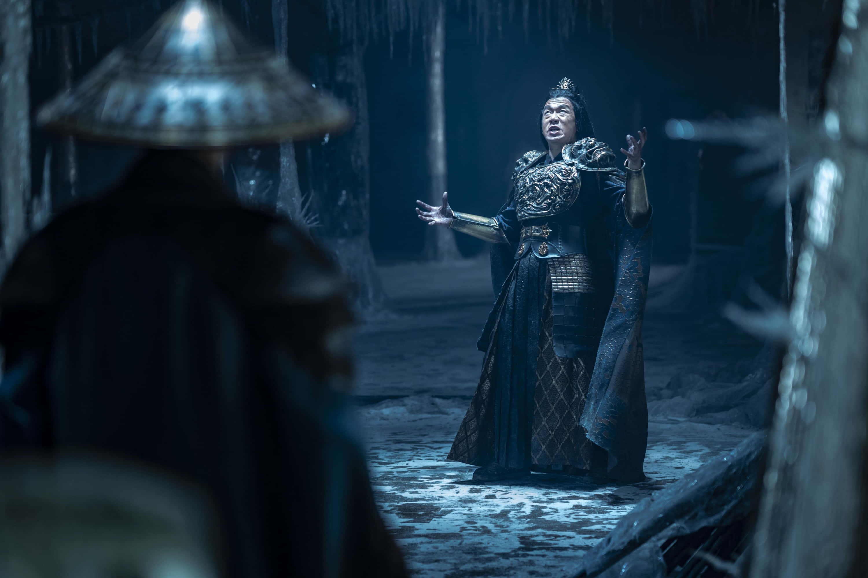 Mortal Kombat Shang Tsung 桌布 壁紙 — 一名身穿華麗盔甲的戰士站在昏暗的冰洞中，在冰冷的藍光下與一個戴兜帽的身影對峙。