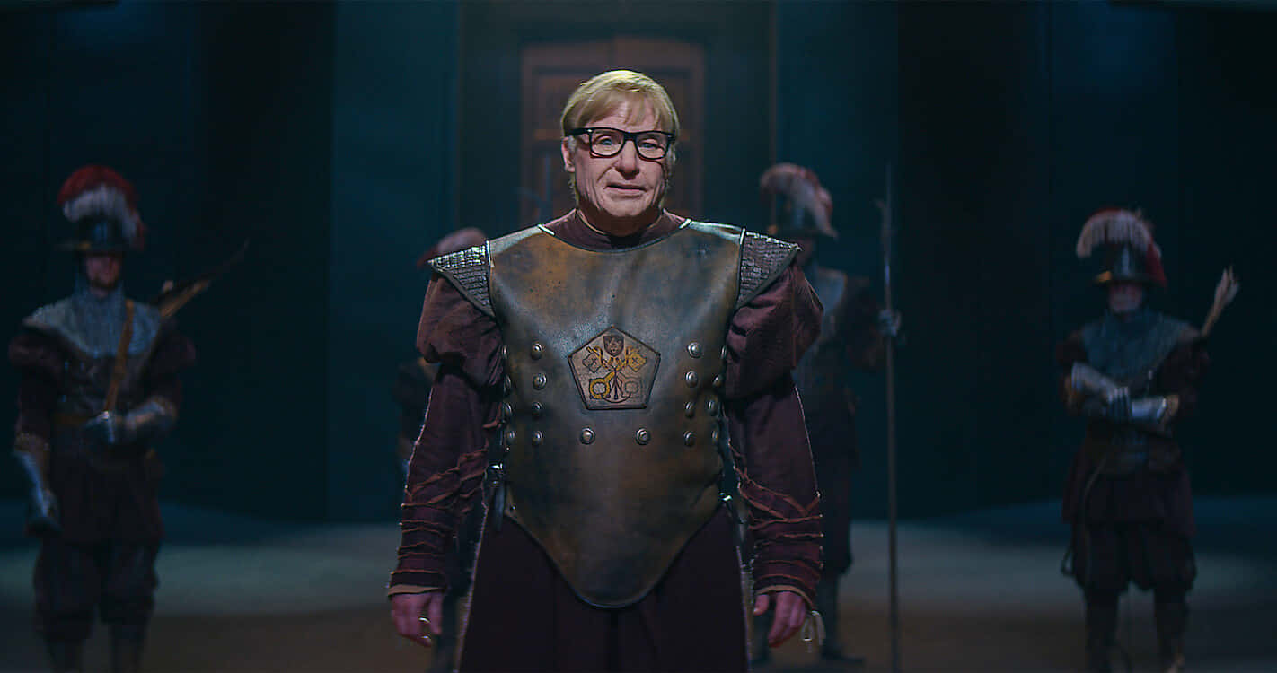 Mike Myers 桌布 壁紙 — 一名身穿華麗盔甲的男人站在舞台中央，兩旁有衛兵，在昏暗的劇場燈光下。