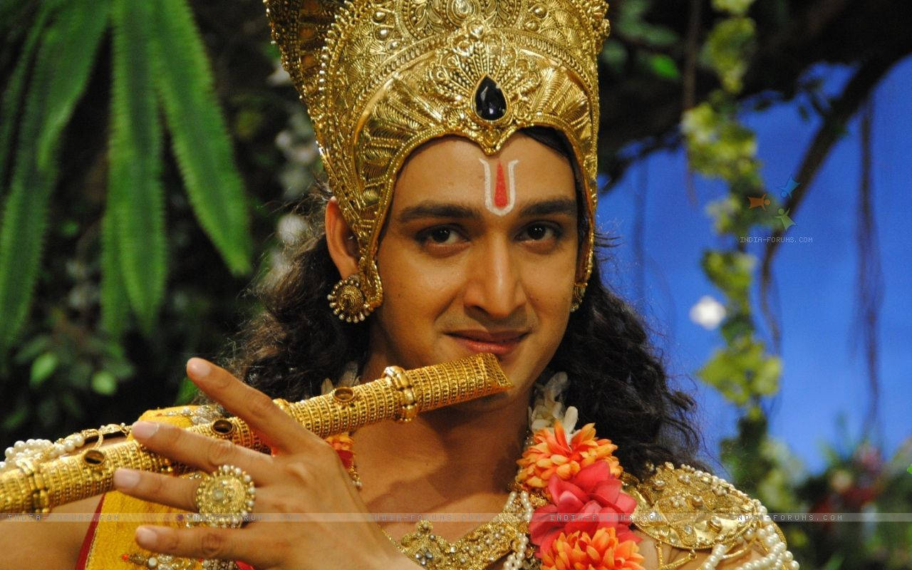 Mahabharat Krishna 桌布 壁紙 — 一名身穿華麗金色神祇服飾的男子在茂密綠葉和藍天下吹奏長笛。