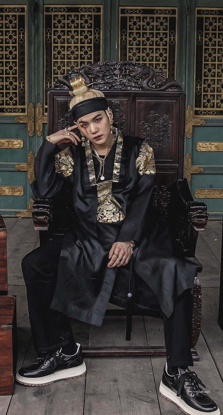 Agust D 桌布 壁紙 — 一名身穿華麗黑金傳統服飾的男子，威嚴地坐在一張雕刻精美的木製王座上，背景是綠色和金色的格子門。