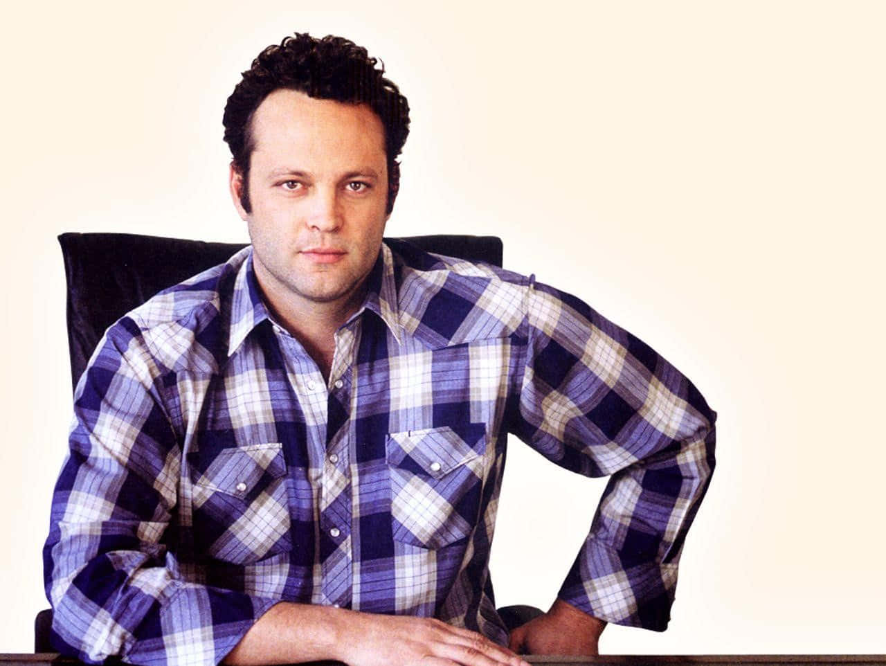 Vince Vaughn 桌布 壁紙 — 一名身穿藍白格仔恤衫的男子自信地坐在辦公桌前，背景是純色米白色。