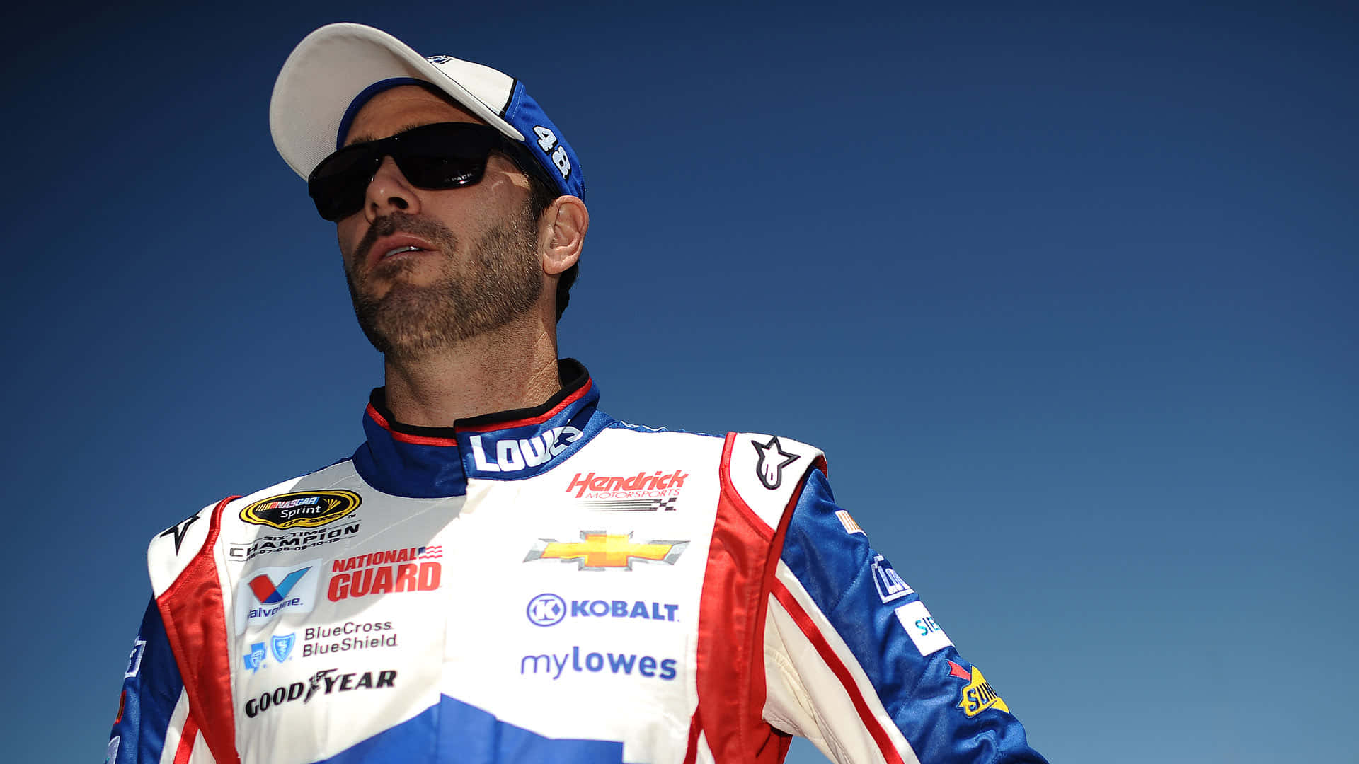 Jimmie Johnson 桌布 壁紙 — 一名身穿藍白紅賽車服、帶有贊助商標誌的NASCAR車手，站在清澈的藍天下。
