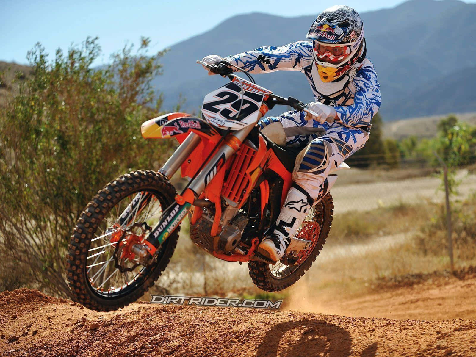Ktm Bike 桌布 壁紙 — 一名身穿藍白色裝備的越野電單車手，駕駛橙色越野電單車，在塵土飛揚的土坡上空飛躍，背景是山脈。