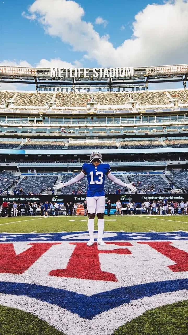 Obj 桌布 壁紙 — 一名身穿藍色13號球衣的美式足球運動員在梅特萊夫體育場的球場上張開雙臂，站在藍天白雲下。
