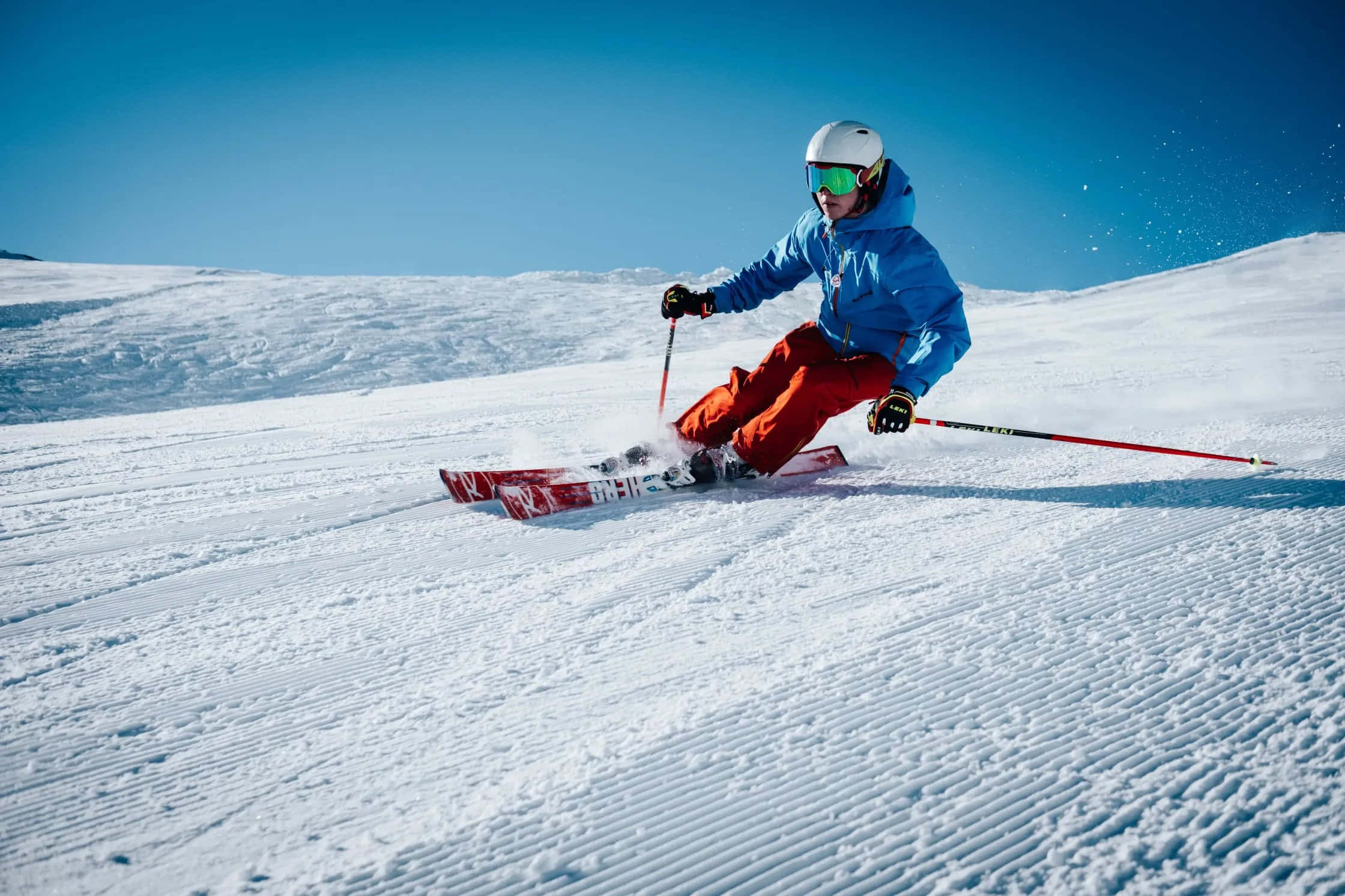 Skiing 桌布 壁紙 — 一名身穿藍色外套和紅色褲子的滑雪者，在晴朗的藍天下沿著修整過的雪坡滑下。