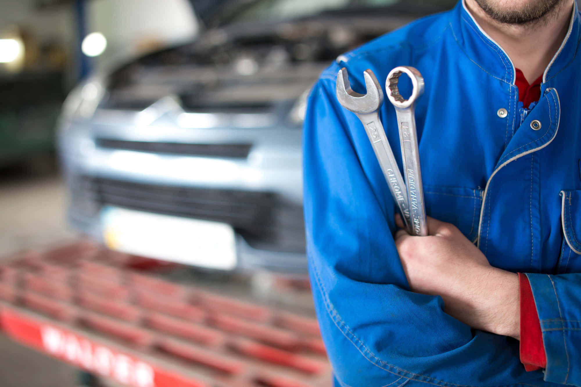 Car Repair 桌布 壁紙 — 一名身穿藍色工作外套的機械師手持兩把扳手，背景模糊中有一輛銀色汽車在紅色升降機上。
