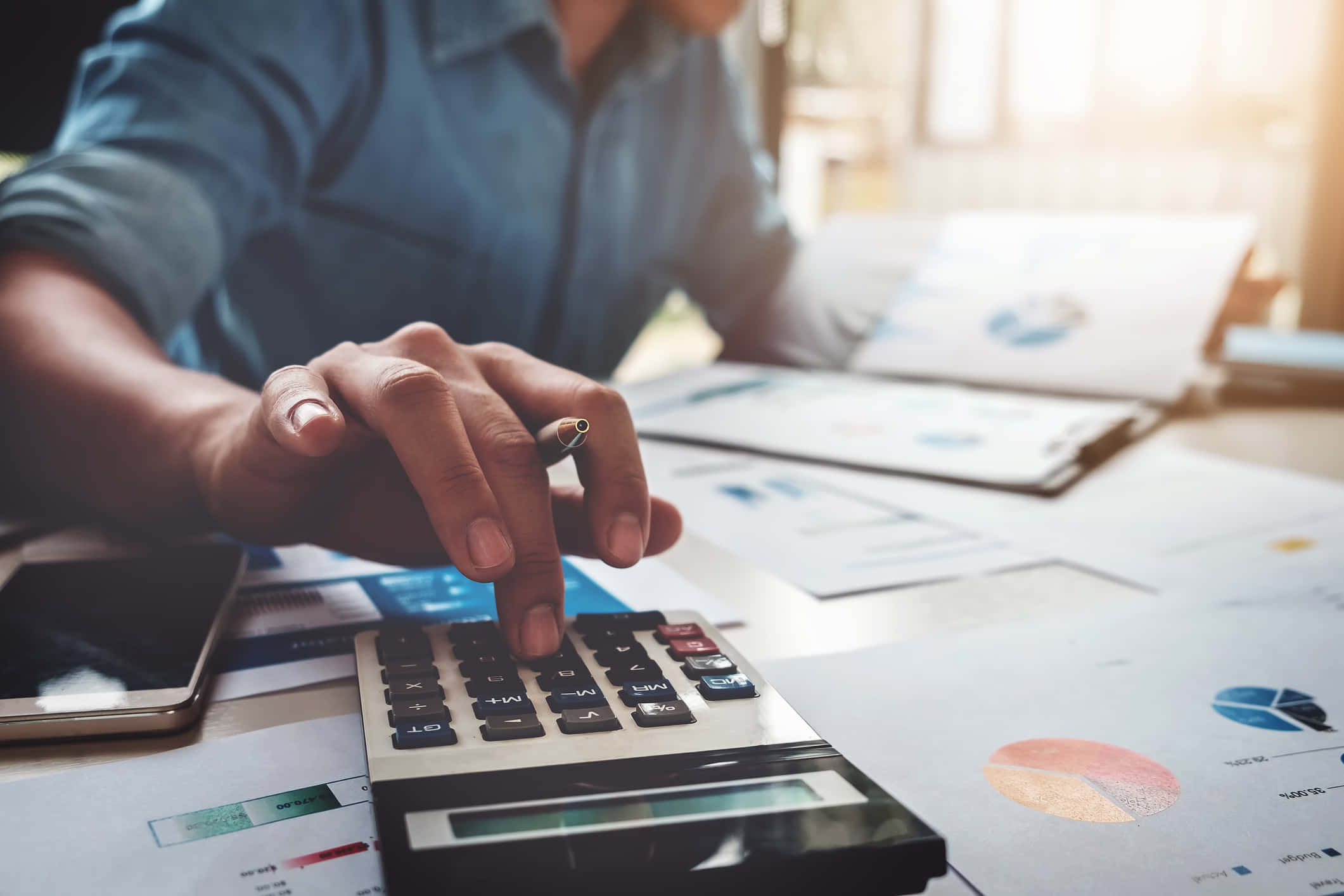 Finance 桌布 壁紙 — 一名身穿藍色恤衫的人士在陽光照射的桌子上使用計算機，周圍是財務圖表和圖形。