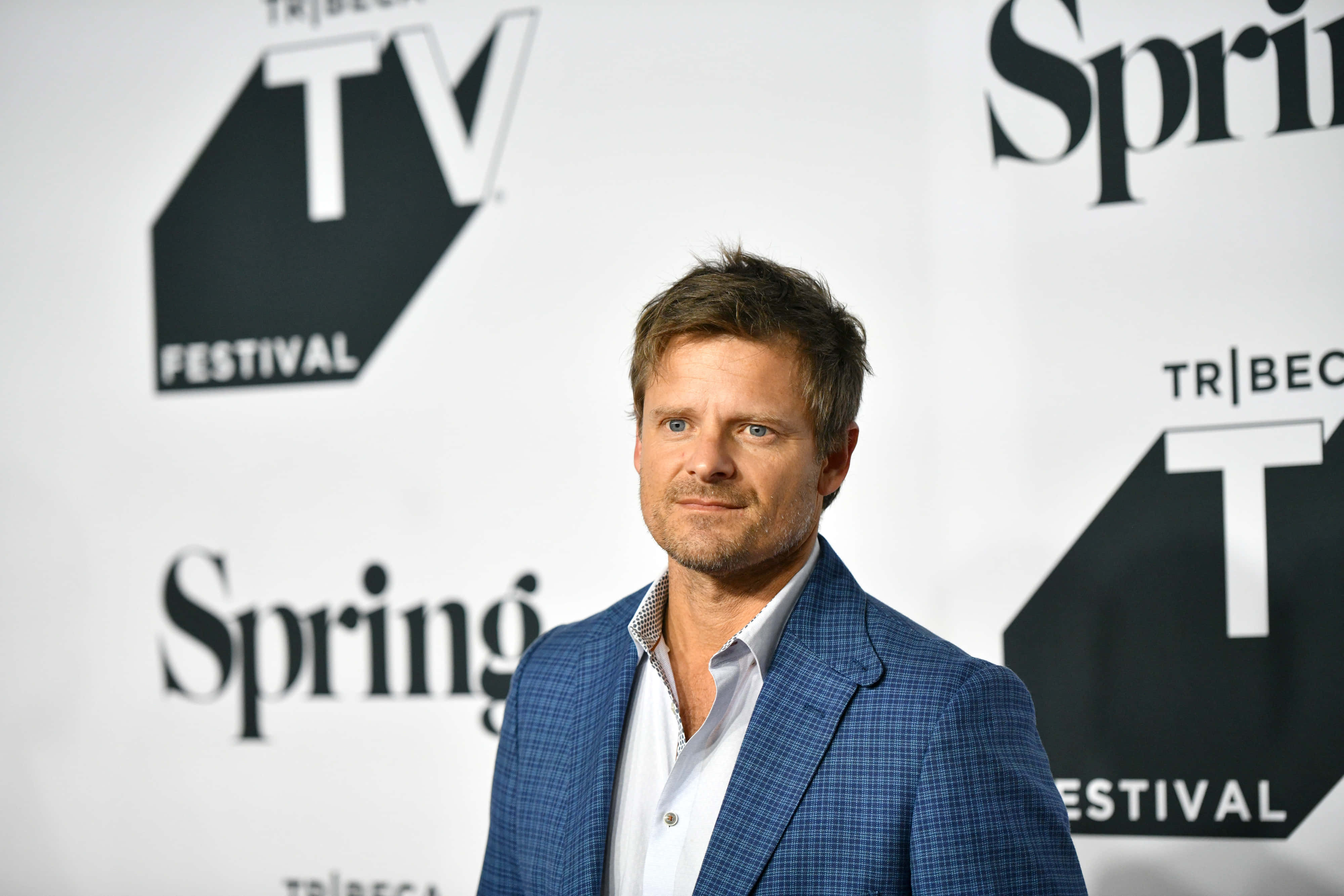 Steve Zahn 桌布 壁紙 — 一名身穿藍色格紋西裝的男子在帶有品牌背景的Tribeca TV Festival活動中自信地站立。