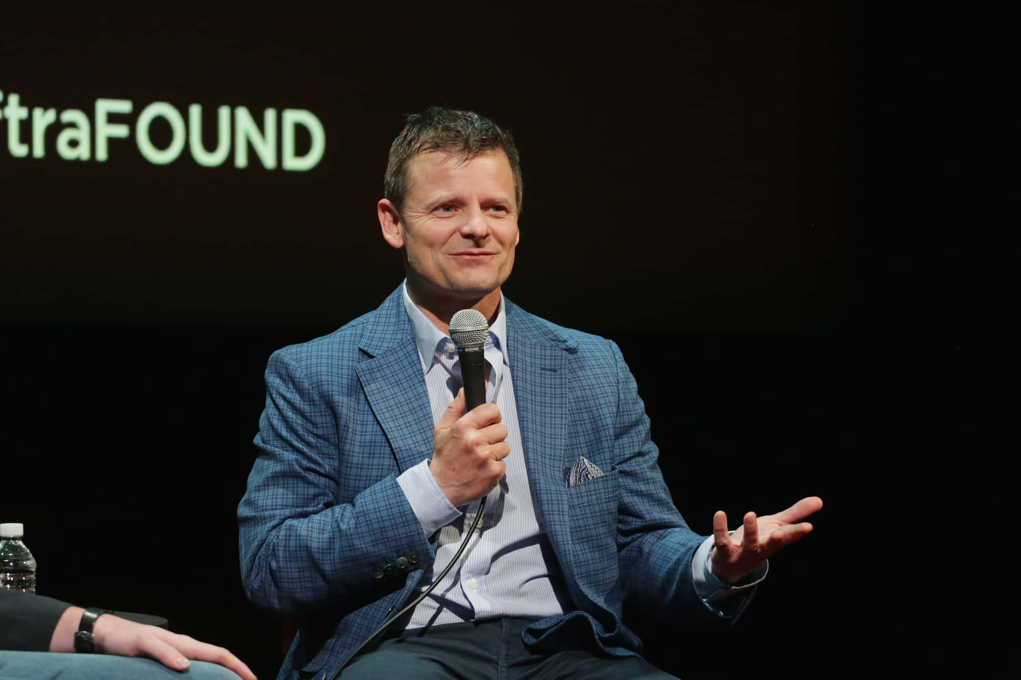 Steve Zahn 桌布 壁紙 — 一名身穿藍色格紋西裝的男子在燈光昏暗的活動中對著麥克風說話，身後投影著「traFOUND」。
