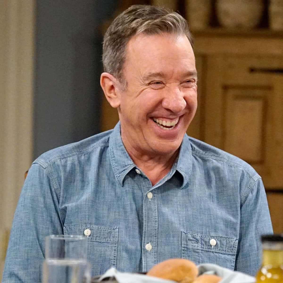 Tim Allen 桌布 壁紙 — 一名身穿藍色牛仔恤衫的男子坐在擺有食物和飲品的餐桌上開懷大笑。