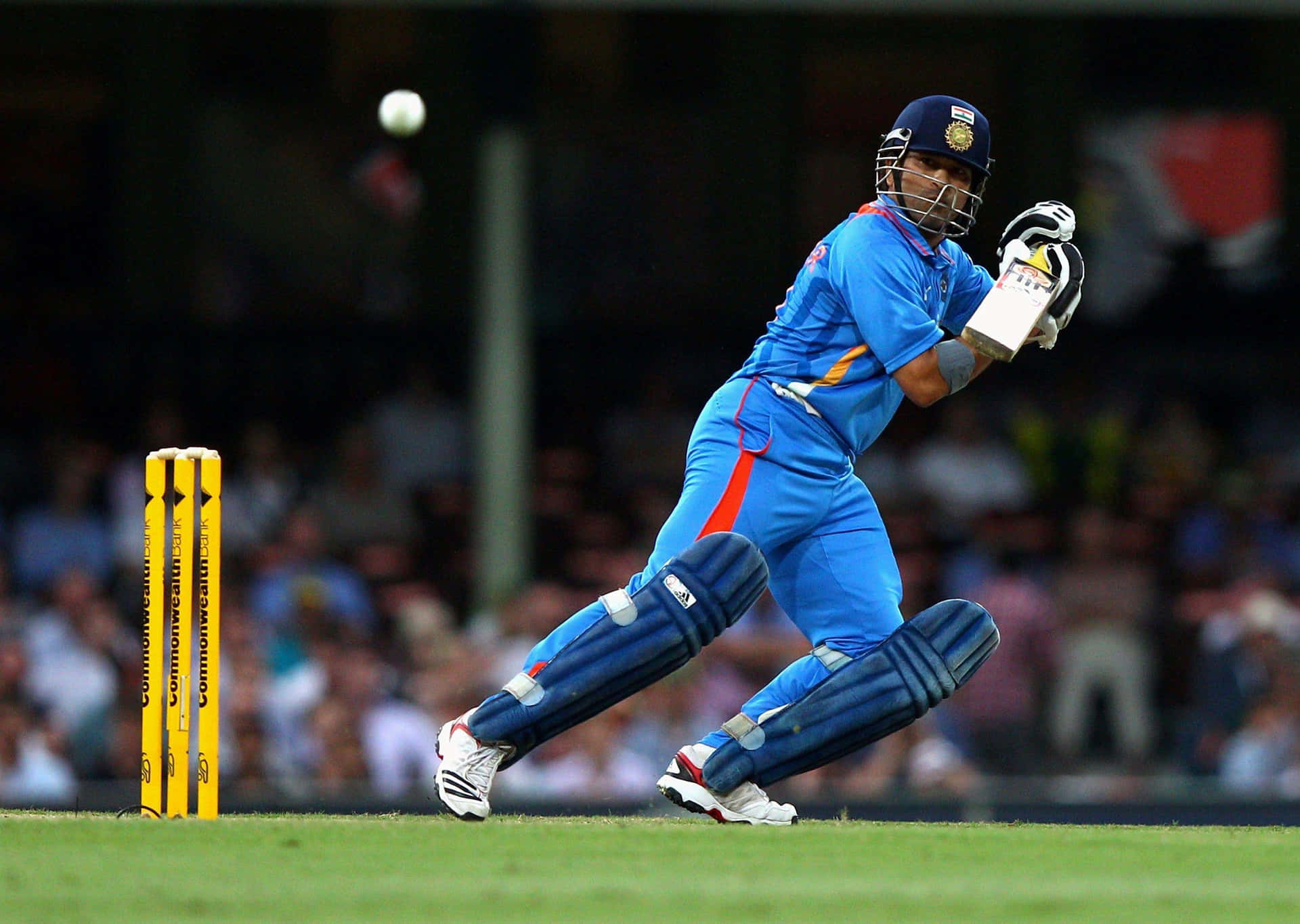 Sachin Tendulkar 桌布 壁紙 — 一名身穿藍色球衣的板球運動員在球場燈光下，於綠色場地上靠近黃色三柱門處揮棒擊打白色球。