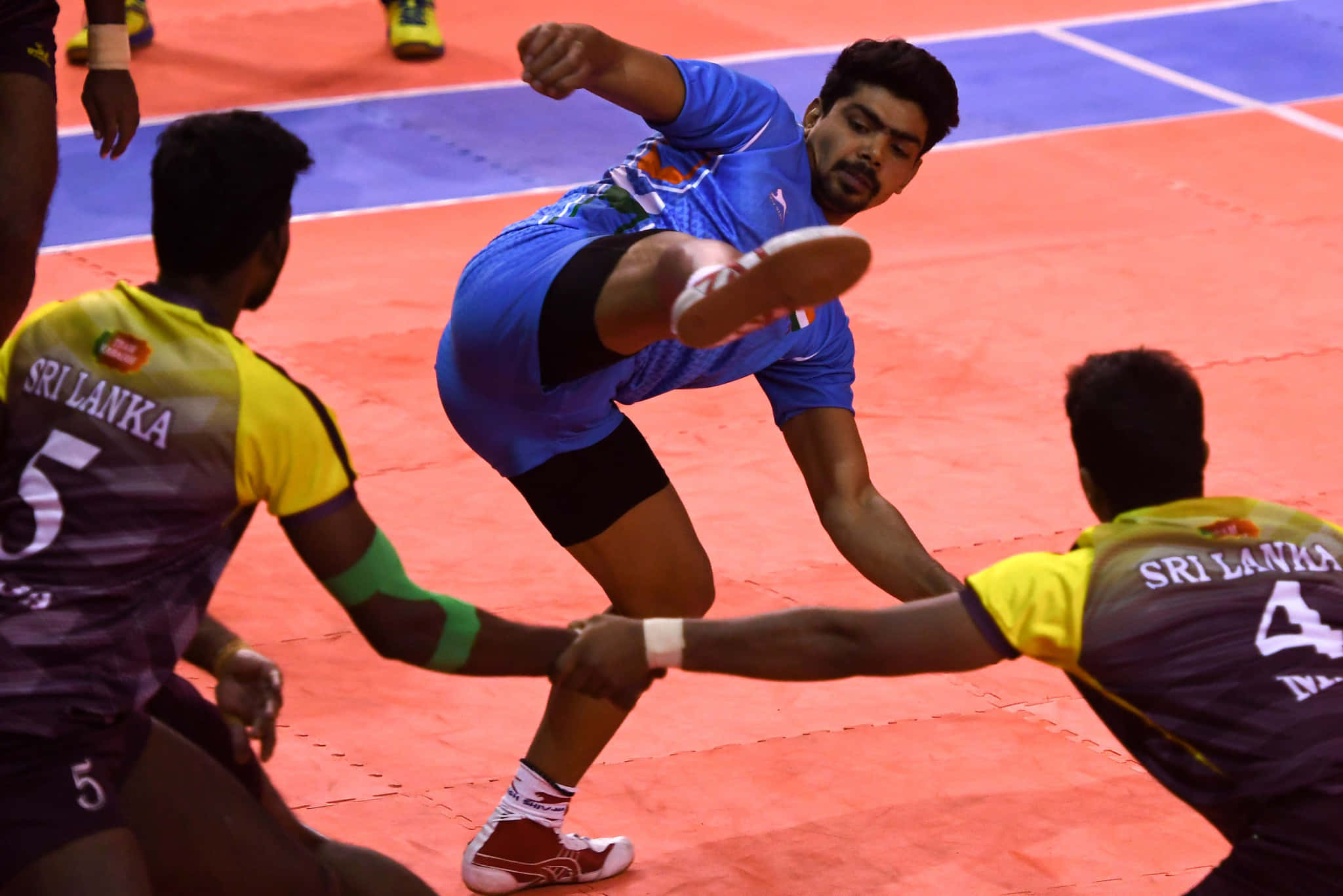 Kabaddi 桌布 壁紙 — 一名身穿藍色球衣的男排球員動態跳起扣球，而身穿黃色和紫色球衣的對手正在防守。