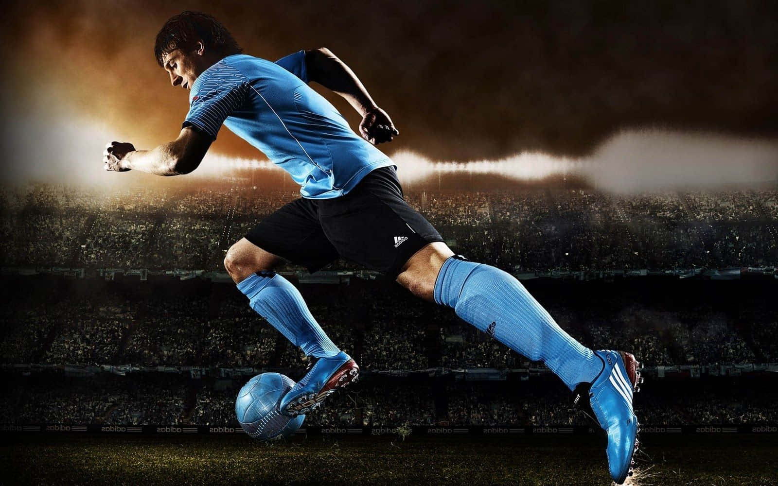 3d Sports 桌布 壁紙 — 一名身穿藍色球衣的足球員在球場燈光下，以動態動作踢球，背景是黑暗且擠滿觀眾的場景。